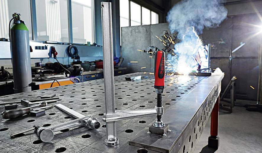 Eine Metallwerkstatt mit Schweißen im Hintergrund und einem Spannwerkzeug auf einem Tisch im Vordergrund.