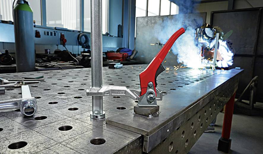 Eine Werkstatt mit einer Metall-Arbeitsfläche, auf der eine Metallklemme und ein Griff zu sehen sind. Im Hintergrund Funken von Schweißarbeiten.