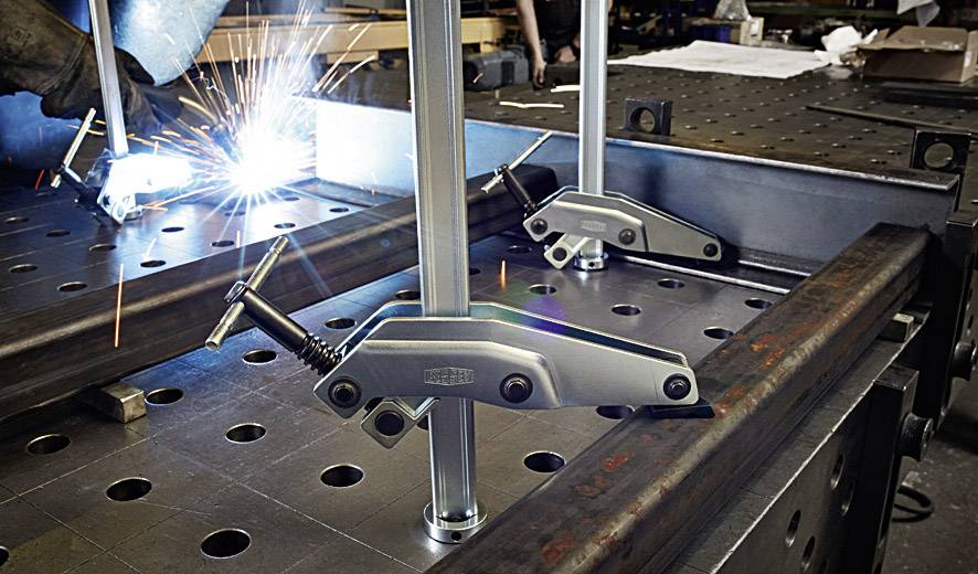 Eine Schweißszene in einer Werkstatt. Funken fliegen, während Metallteile mit Klemmen auf einem Werkbank zusammengefügt werden.