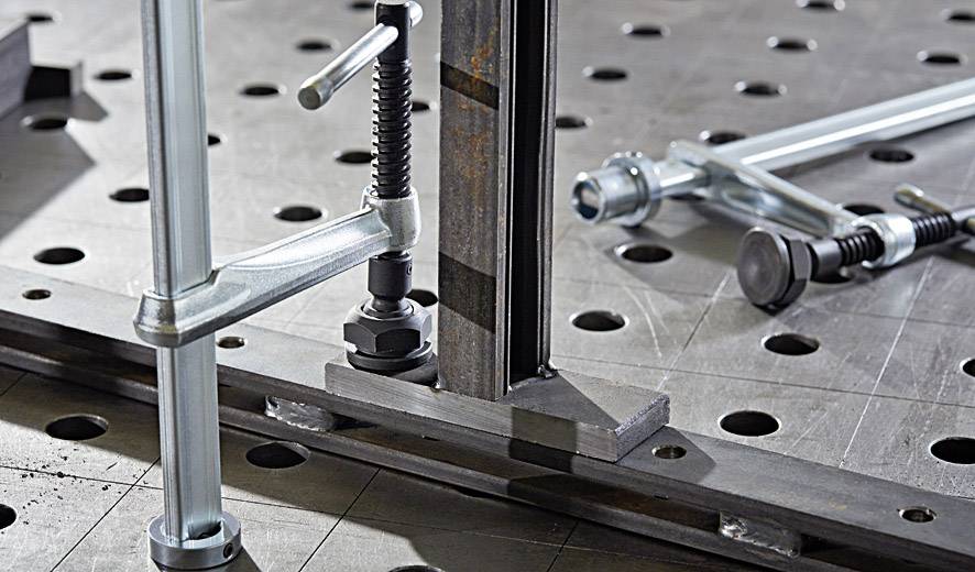 Metallische Klemmen und Schrauben sichern eine Stahlplatte mit Lochmuster auf einem Werkbanktisch, gezeigt in Nahaufnahme.