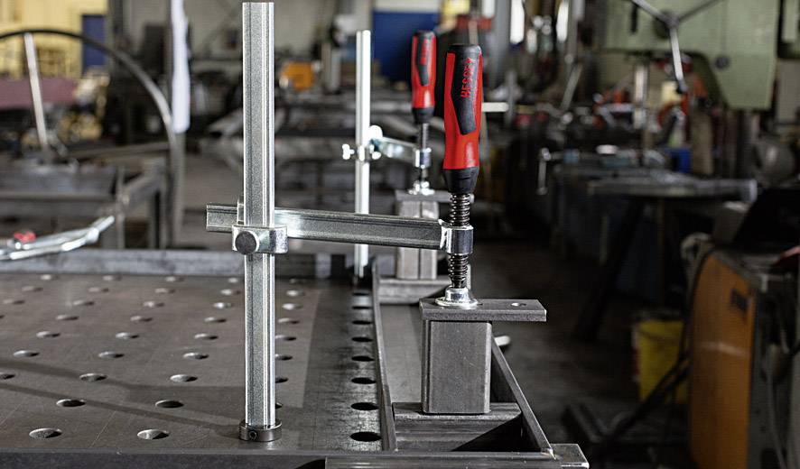 Ein Metalltisch in einer Werkstatt mit mehreren Schraubzwingen, die Metallteile fixieren. Hintergrund zeigt unscharfe Werkzeuge und Maschinen.