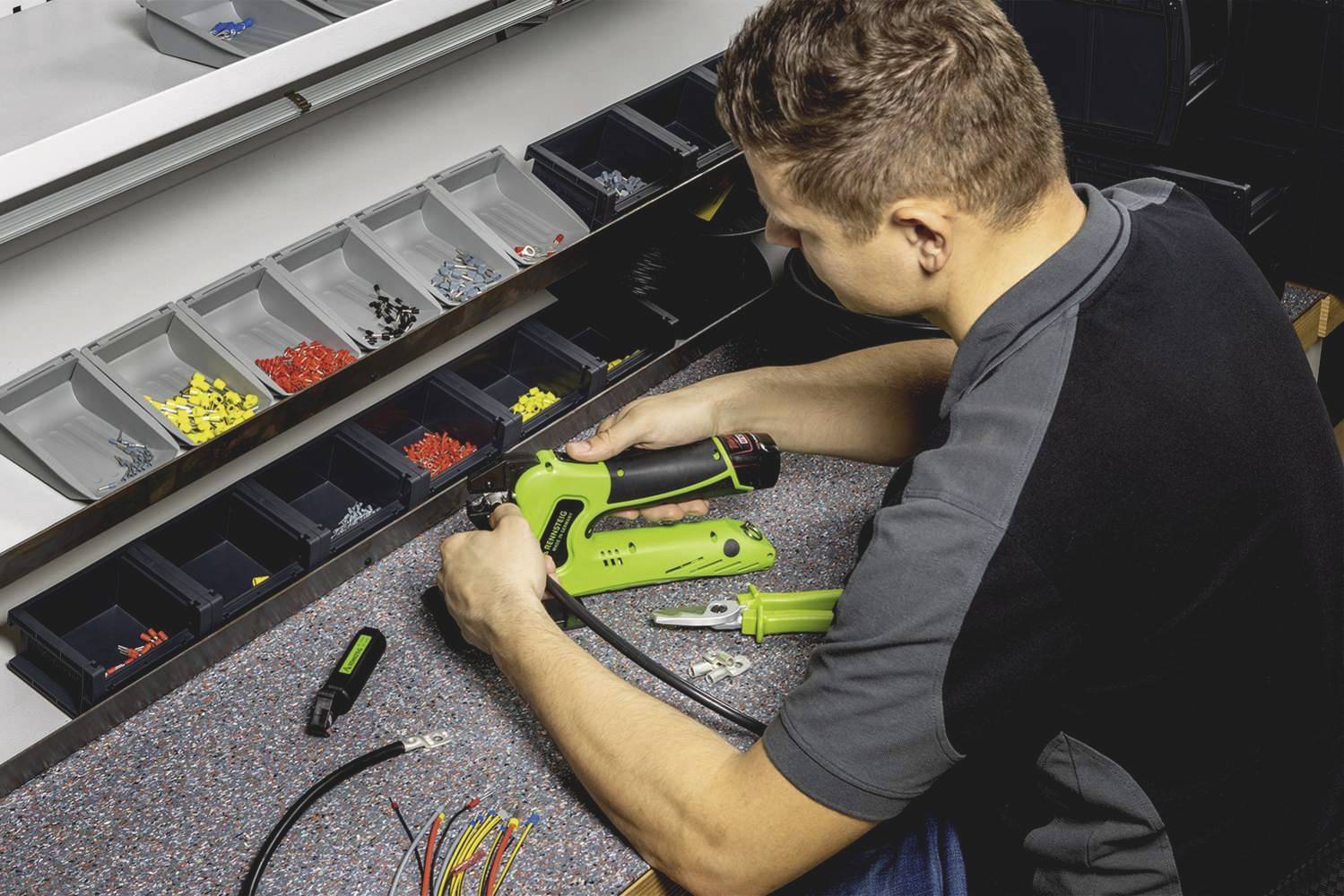 Ein Mann an einem Werkbanksortiment arbeitet mit einem grünen Crimpwerkzeug an Kabeln und Steckverbindern, umgeben von Schraubensortimenten.