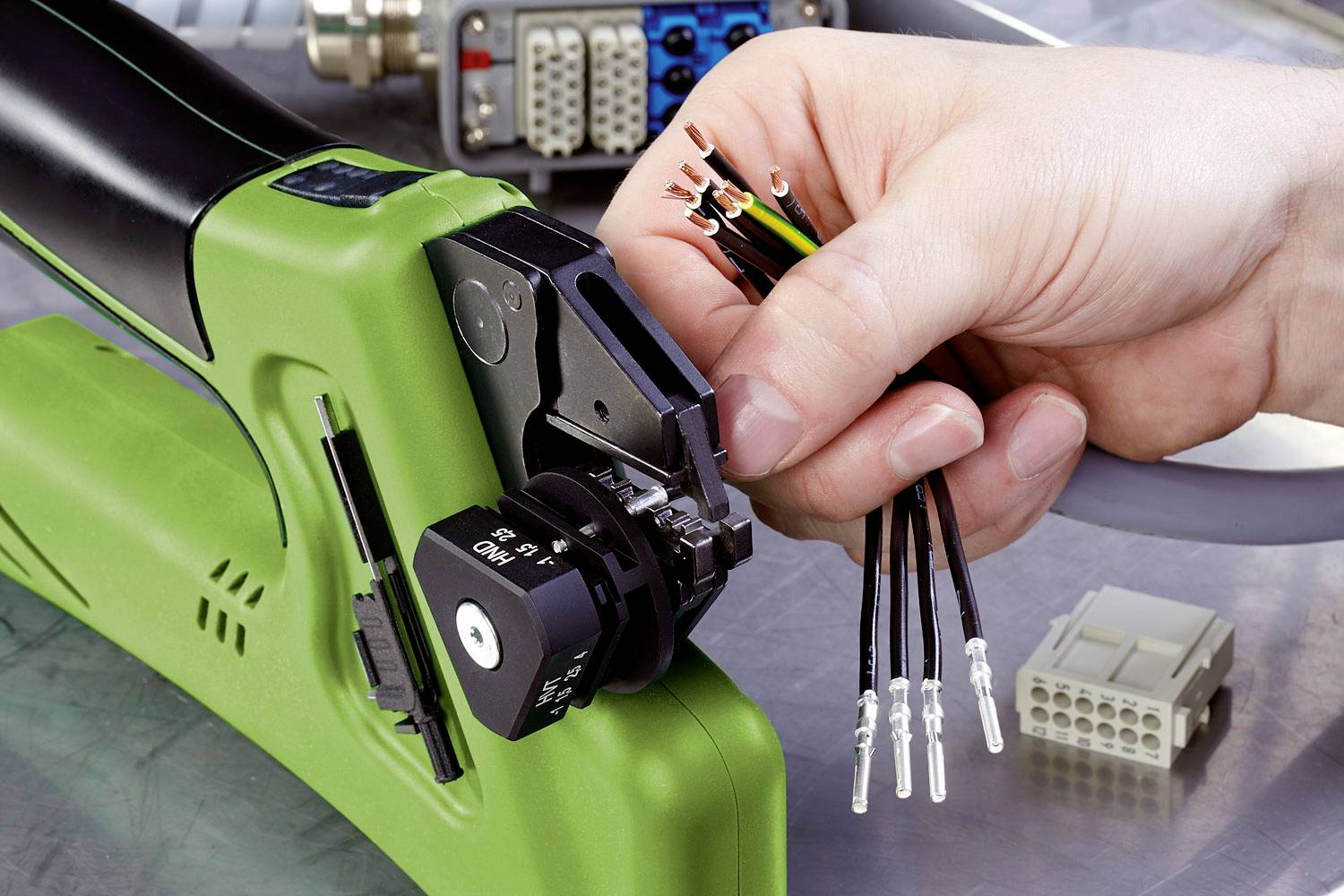 Eine Hand hält Kabel, die in eine grüne Crimpzange eingespannt werden. Im Hintergrund sind verschiedene Steckverbinder sichtbar.