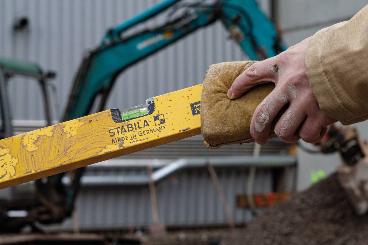 Eine Hand hält eine gelbe Wasserwaage auf einer Baustelle. Im Hintergrund ist ein unscharfer Bagger zu sehen.