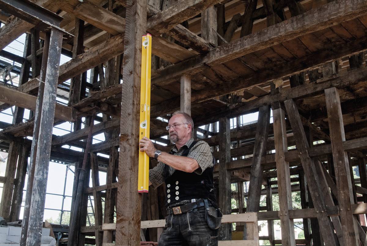 Ein Mann in Arbeitskleidung verwendet eine Wasserwaage, um einen Holzrahmen in einem unfertigen Holzgebäude zu messen.