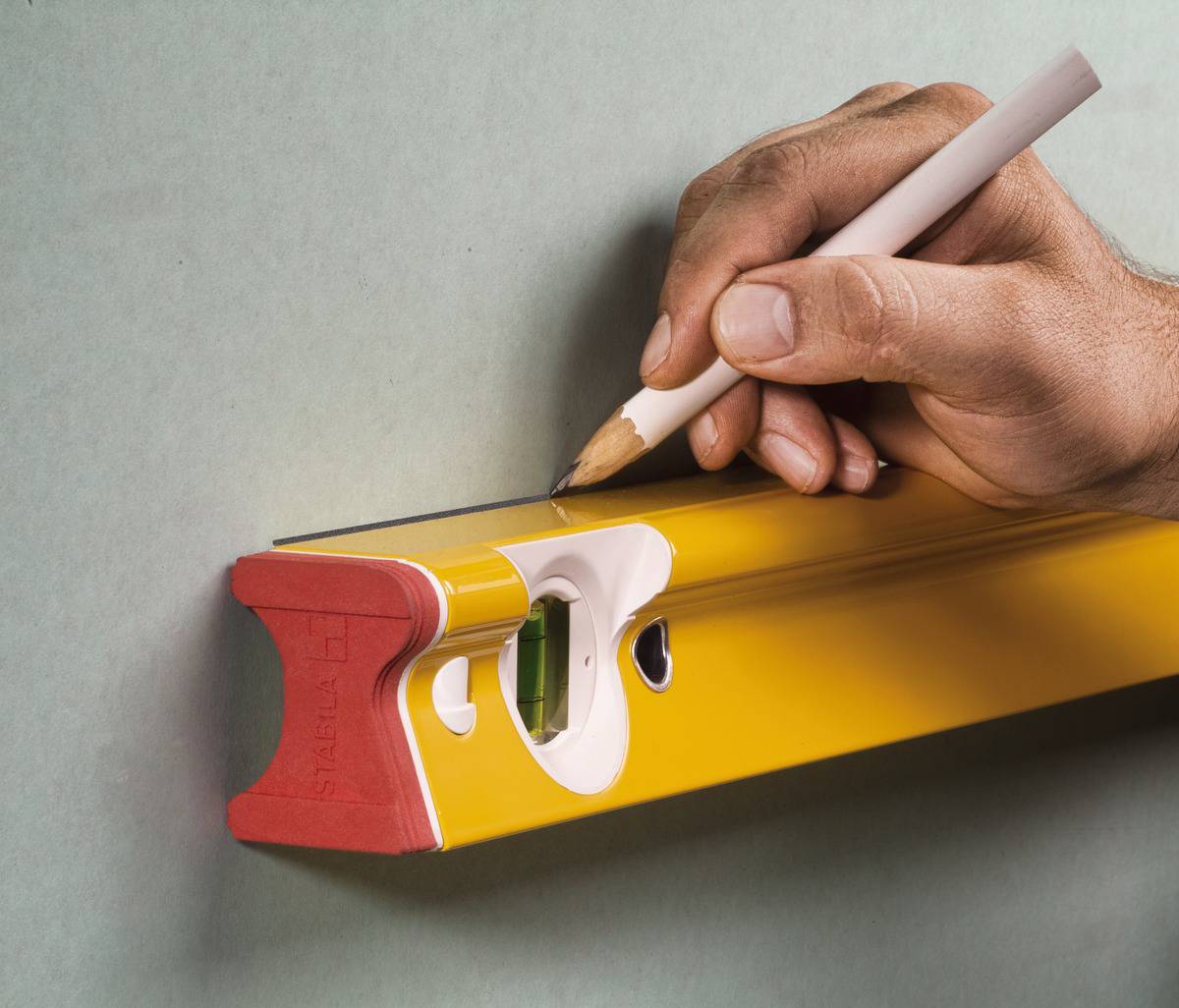 Eine Hand hält einen Bleistift und zeichnet eine Linie entlang einer gelben Wasserwaage, die an einer grünen Wand gehalten wird.