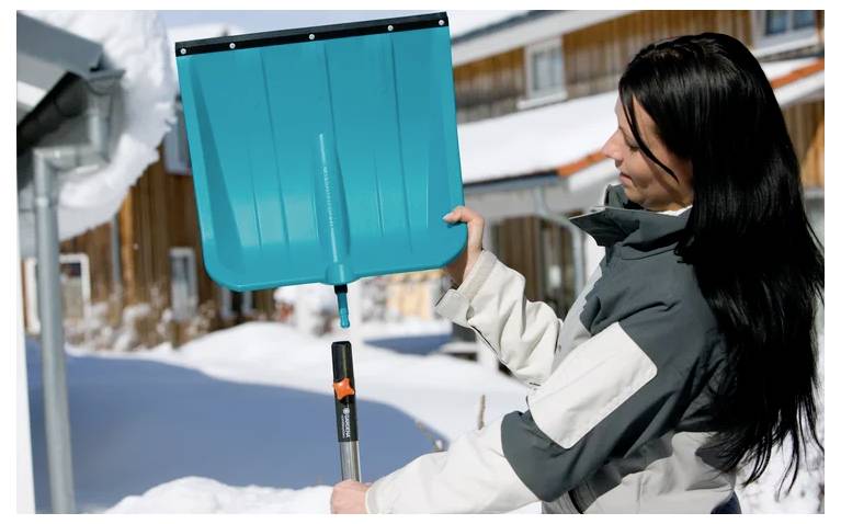 Eine Person in Winterkleidung hält eine Schneeschaufel draußen, mit schneebedeckten Gebäuden im Hintergrund sichtbar.