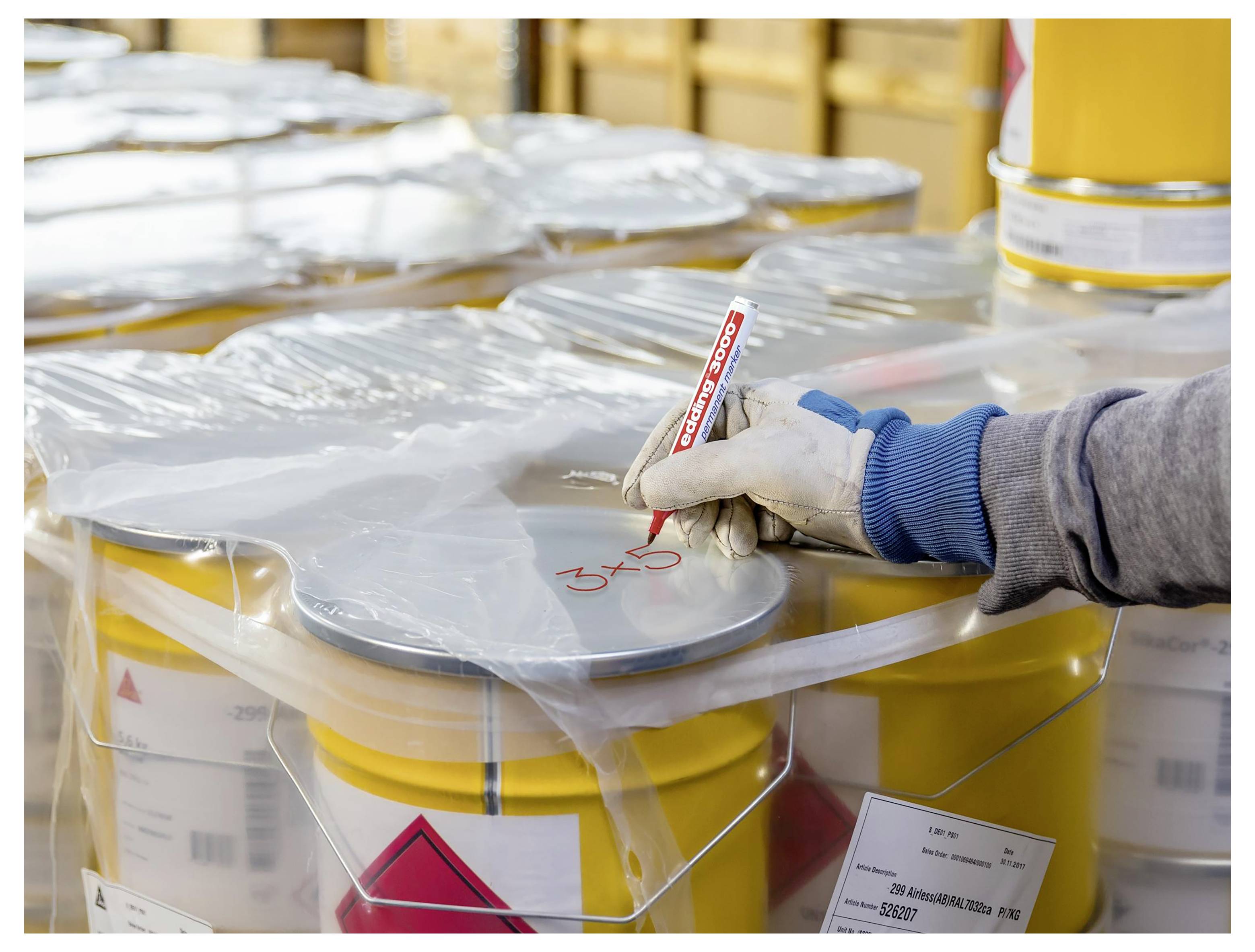 Arbeiter mit Handschuhen beschriftet ein Behältnis mit Gefahrstoffen mit einem Marker in einem Lager