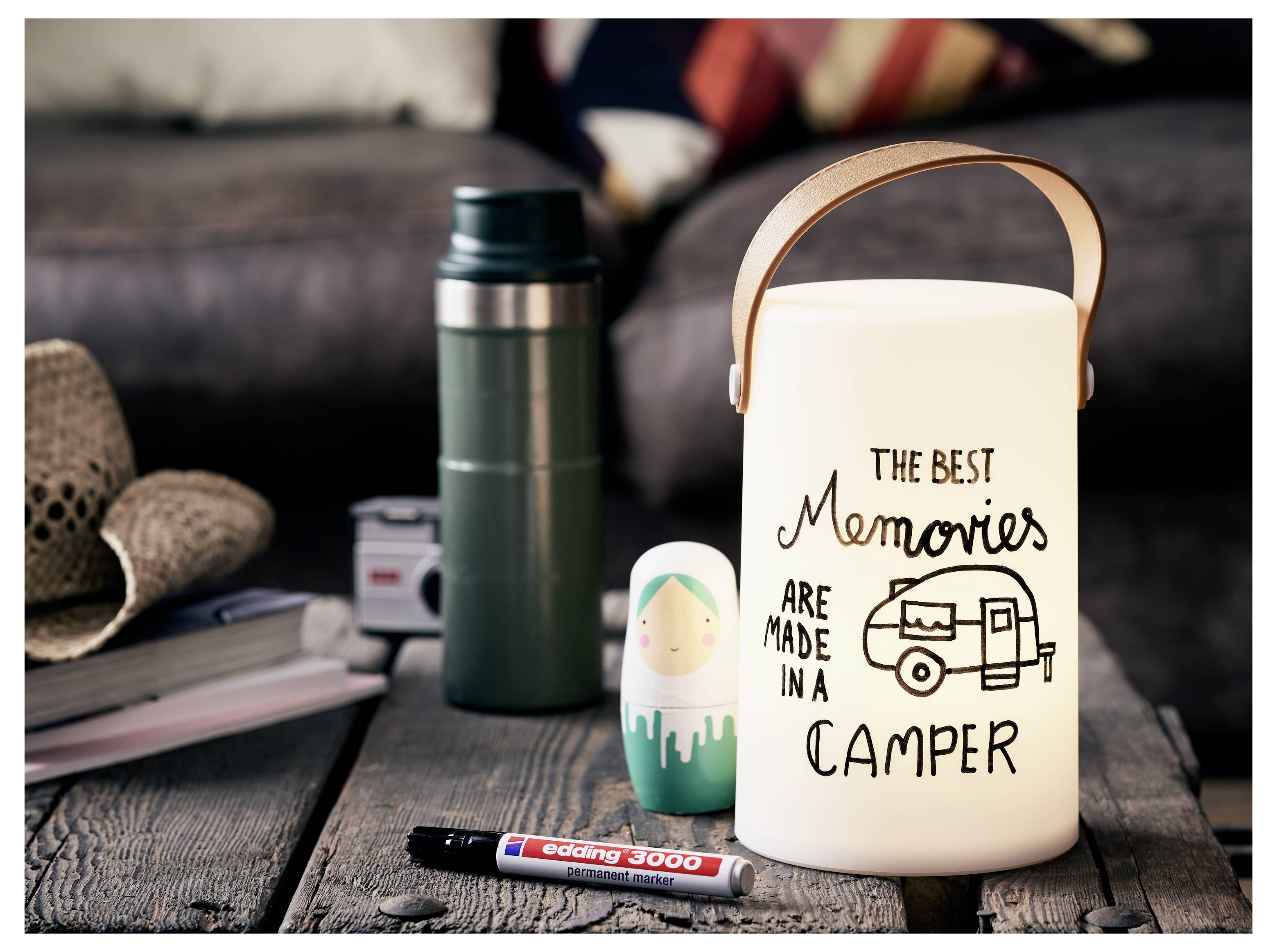 Eine dekorierte Laterne mit der Aufschrift „Die besten Erinnerungen entstehen im Wohnmobil" steht auf einem rustikalen Tisch neben einer Thermoskanne, einem Marker und einer kleinen Wohnmobil-Figur.