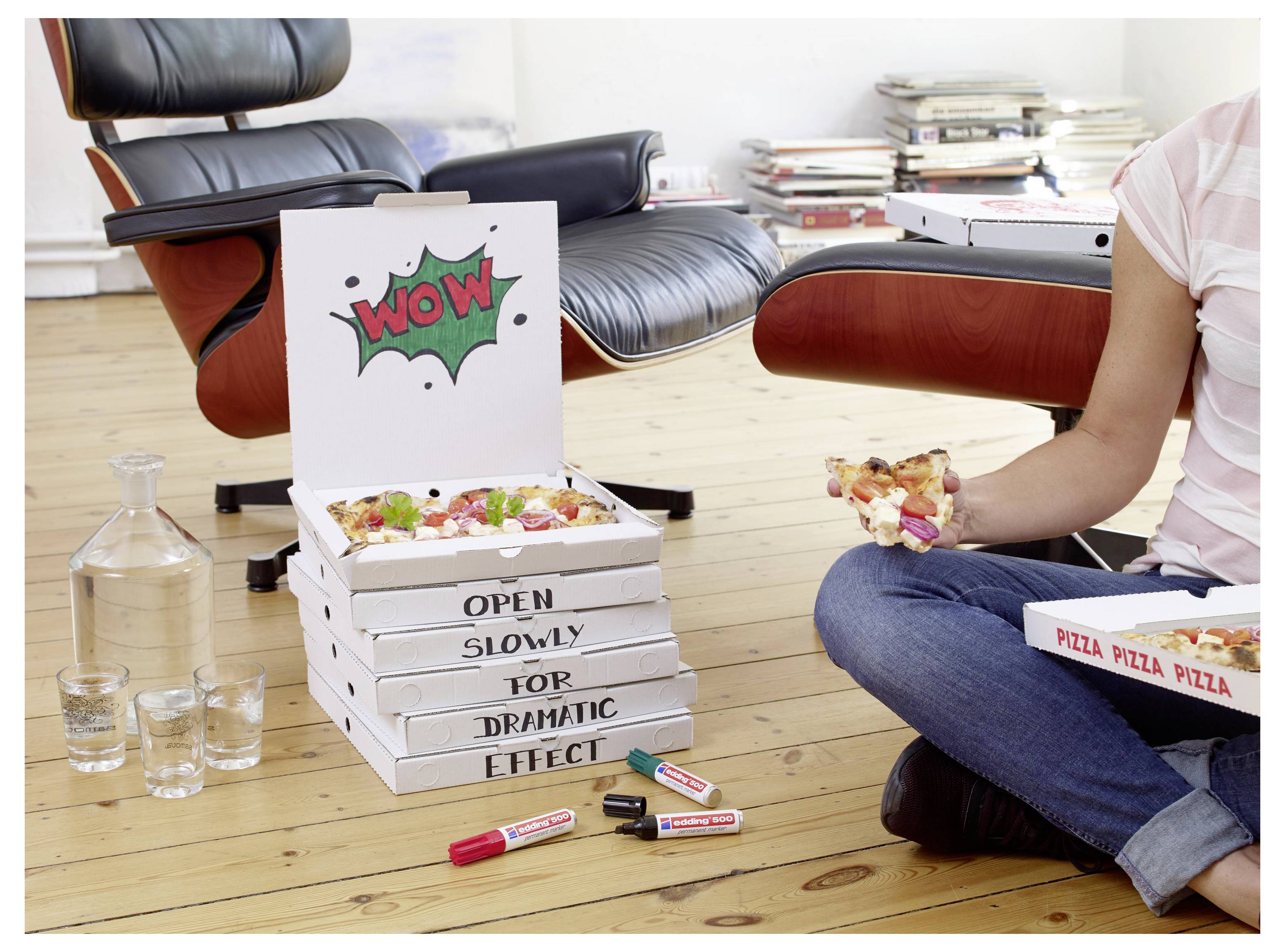 Person sitzt auf dem Boden und isst Pizza neben einem Stapel von Pizzakartons mit der Aufschrift „SLOWLY OPEN FOR DRAMATIC EFFECT" und einem „WOW"-Schild.