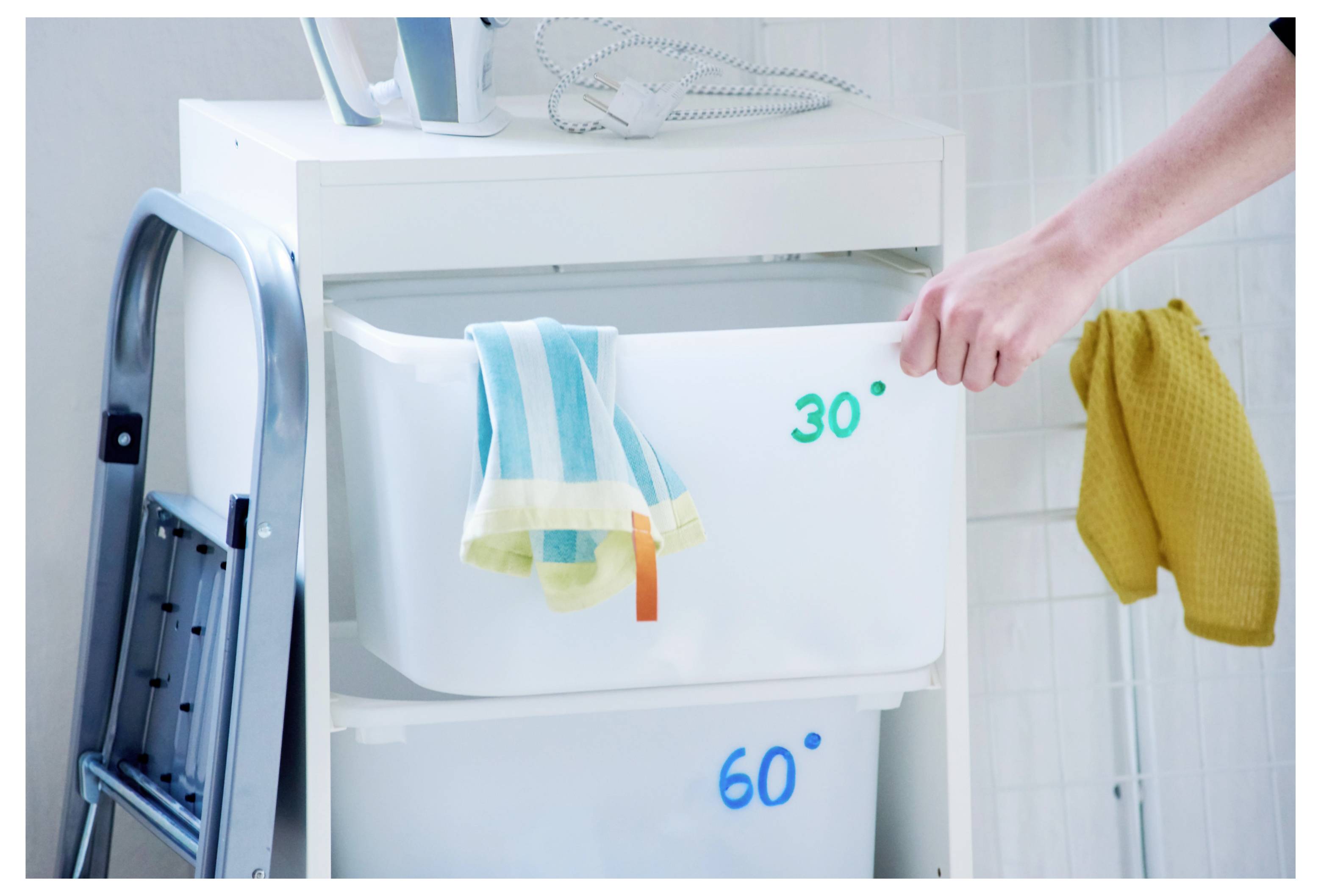 Hand öffnet eine weiße Wäschelade mit der Beschriftung „30°" über einer weiteren mit der Beschriftung „60°", daneben liegen zusammengefaltete Handtücher und Reinigungstücher neben einem Mopp und einem gelben Handtuch.