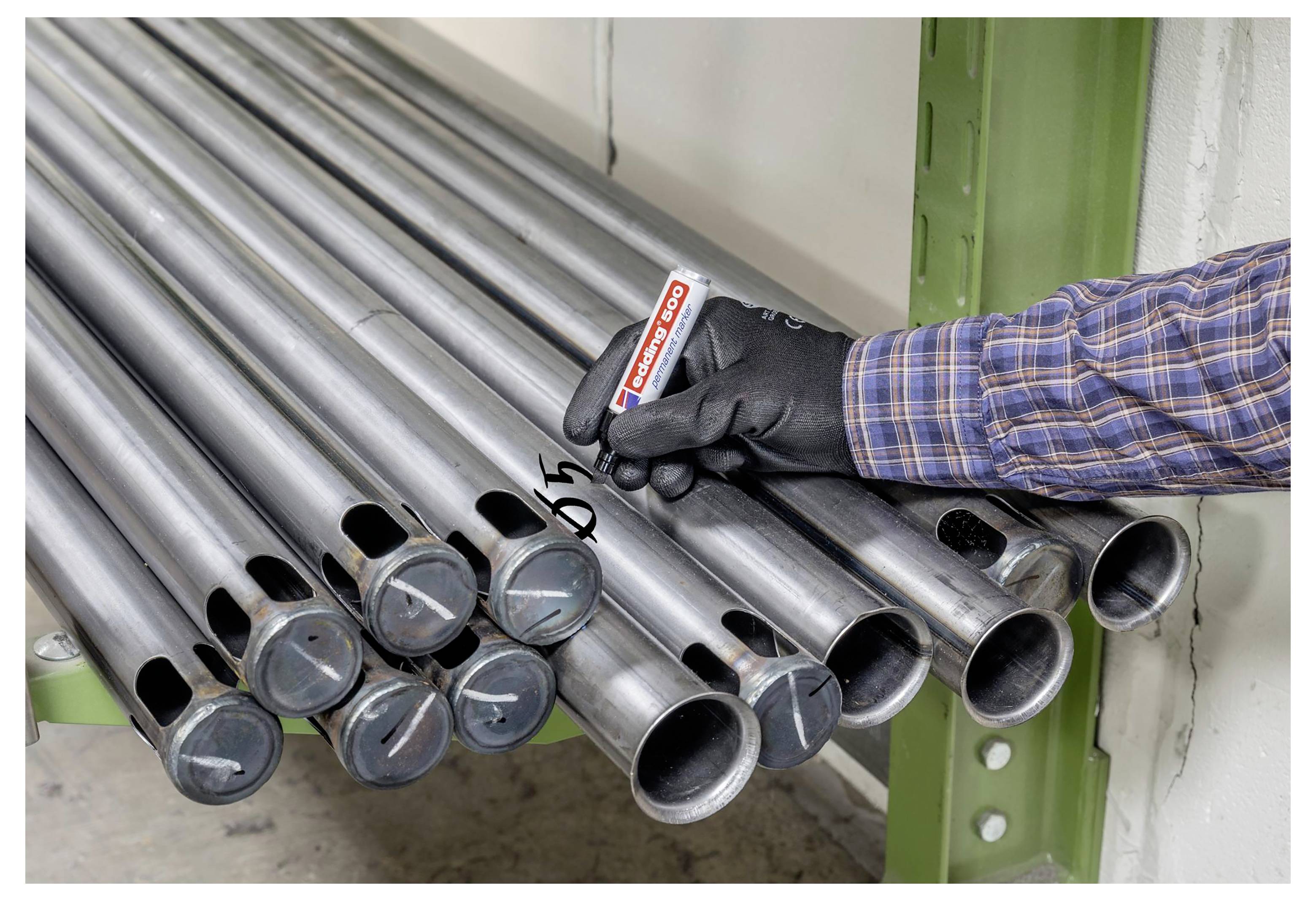 Ein Arbeiter mit Handschuhen markiert eines von mehreren gestapelten Metallrohren mit einem schwarzen Marker in einem industriellen Lagerbereich.