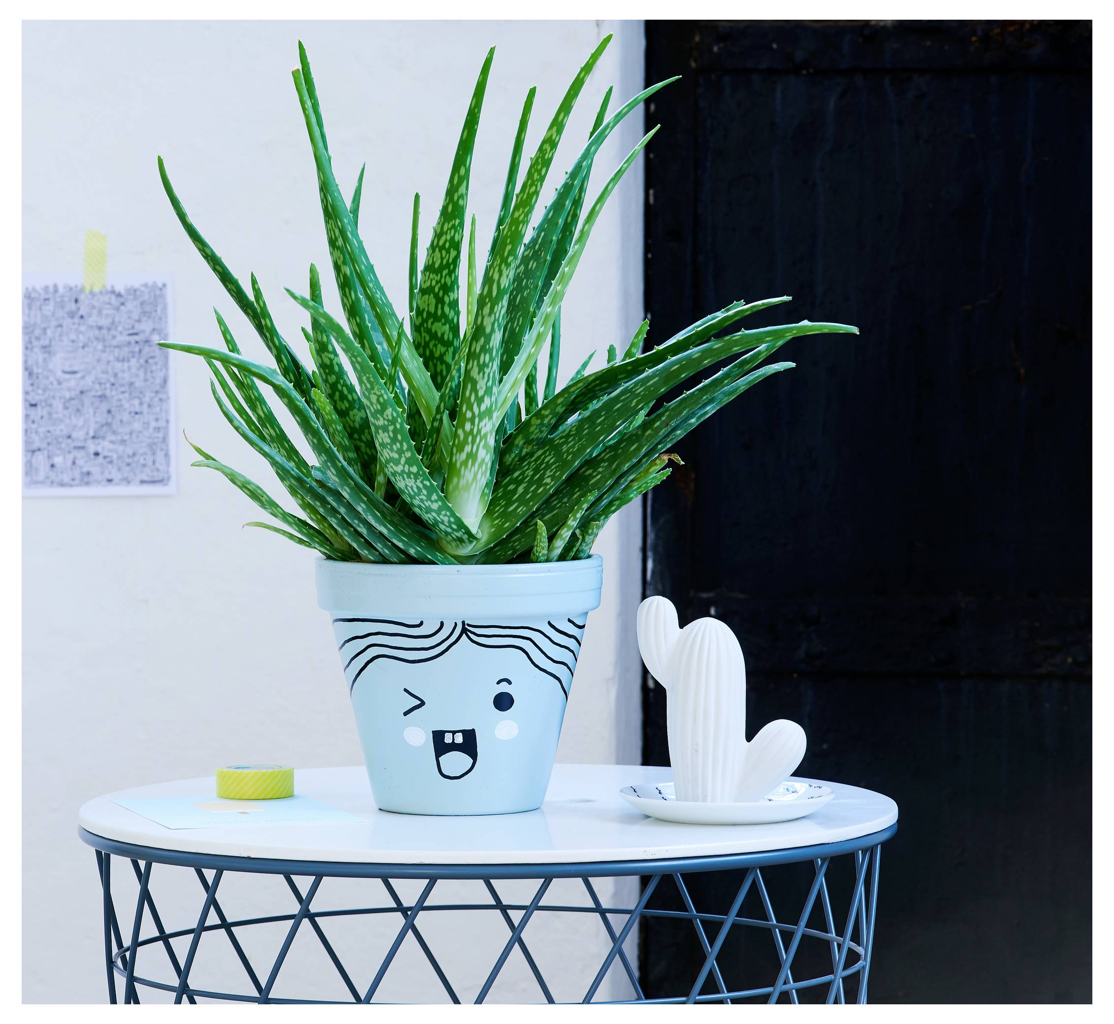 Eine getopfte Aloe-Pflanze in einem blauen Topf mit einem Cartoon-Gesicht sitzt auf einem kleinen runden Tisch neben einer weißen Kaktus-förmigen Dekorfigur.