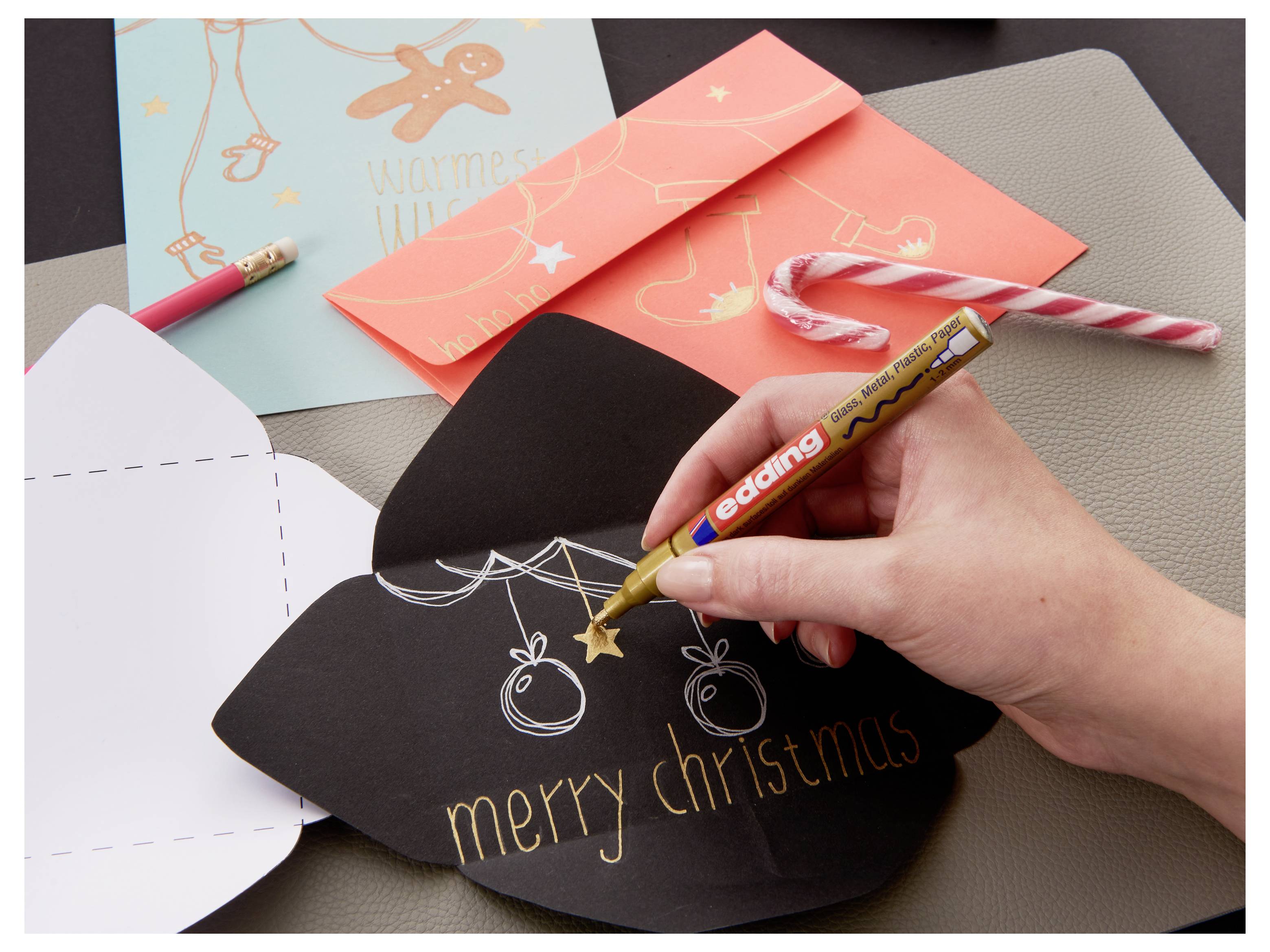 Eine schwarze Weihnachtskarte mit einem goldenen Marker von Hand verzieren, mit anderen festlichen Karten, Buntstiften und Zuckerstange auf dem Tisch.