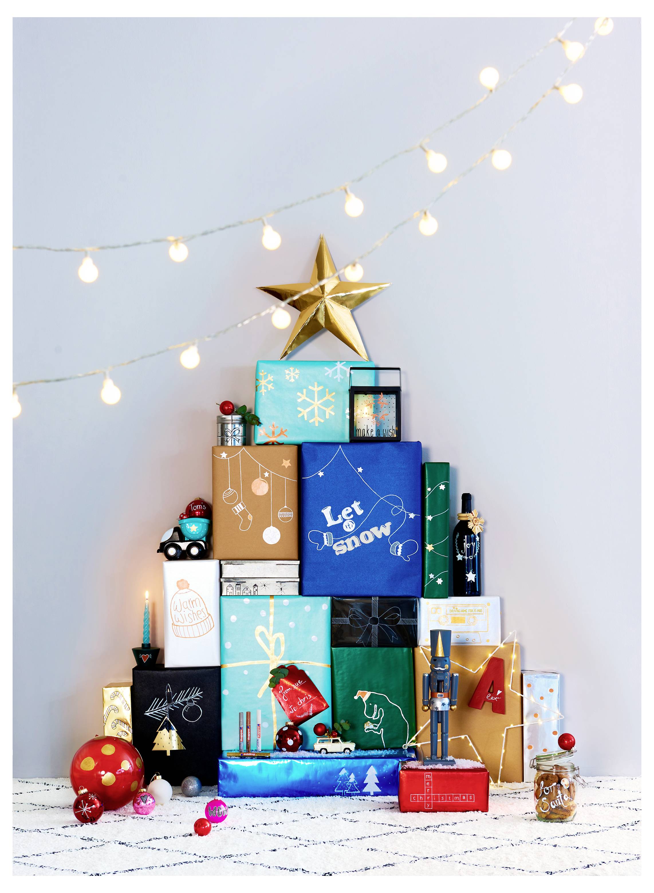 Ein Stapel strahlend verpackter Weihnachtsgeschenke mit einem goldenen Stern auf der Spitze, dekoriert mit Lichterketten und Weihnachtsornaments auf einem weißen Boden.