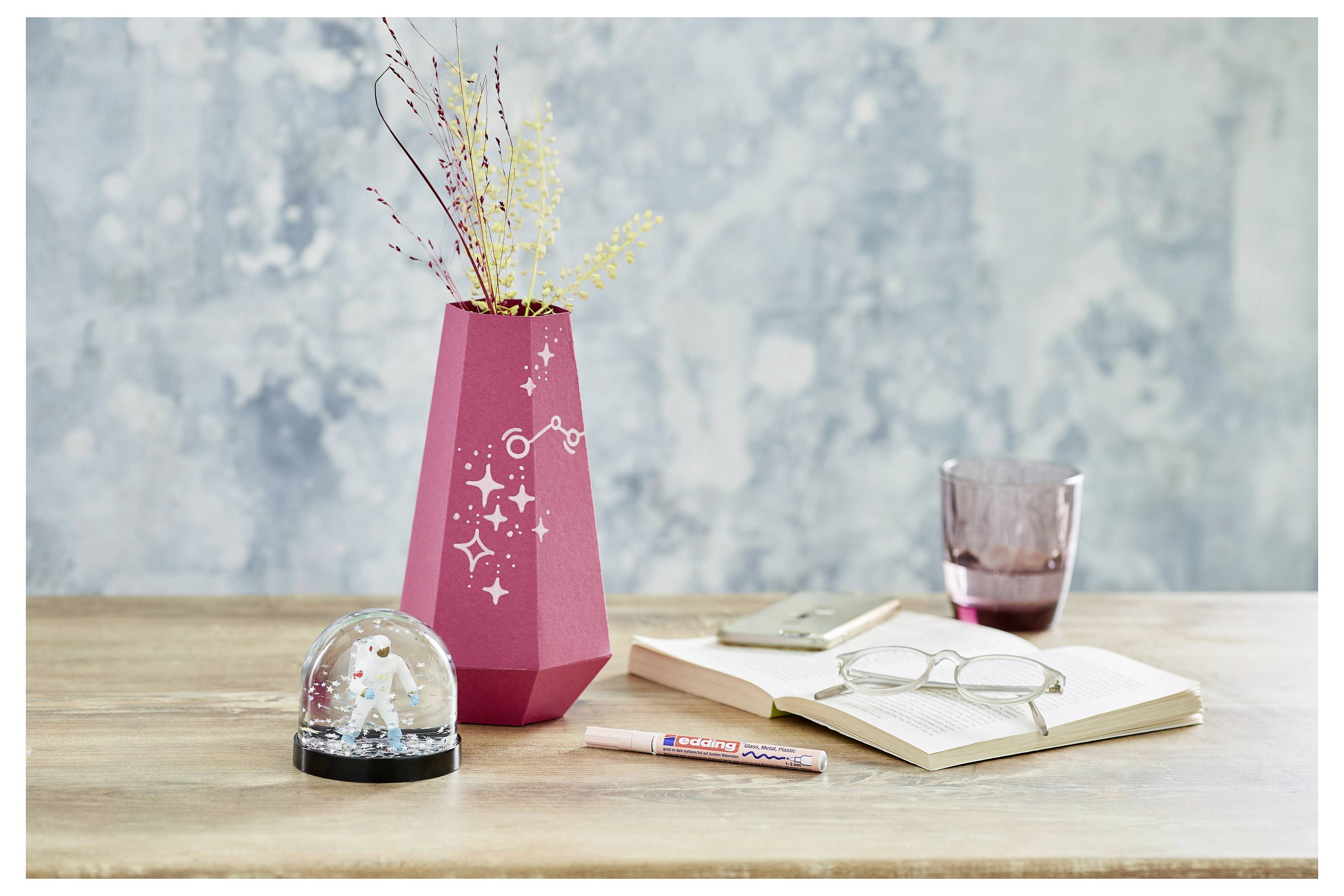 Schreibtischszene mit rosa Vase mit getrockneten Blumen, Schneekugel, geöffnetem Notizbuch mit Brille, Bleistift, Telefon und Glastasse auf einem Holztisch.
