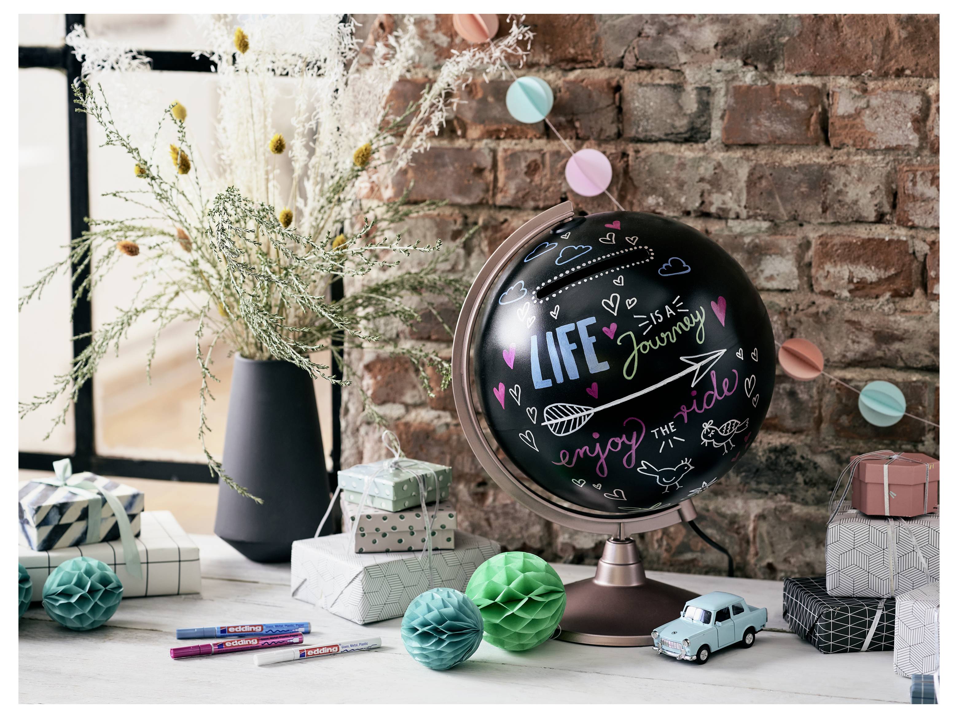 Dekorierter Taffelglobus auf einem Ständer mit bunten Gekritzel und den Worten „LIFE journey enjoy the ride", umgeben von Geschenkboxen, Blumen, Stiften und Spielzeugautos auf einem Tisch.