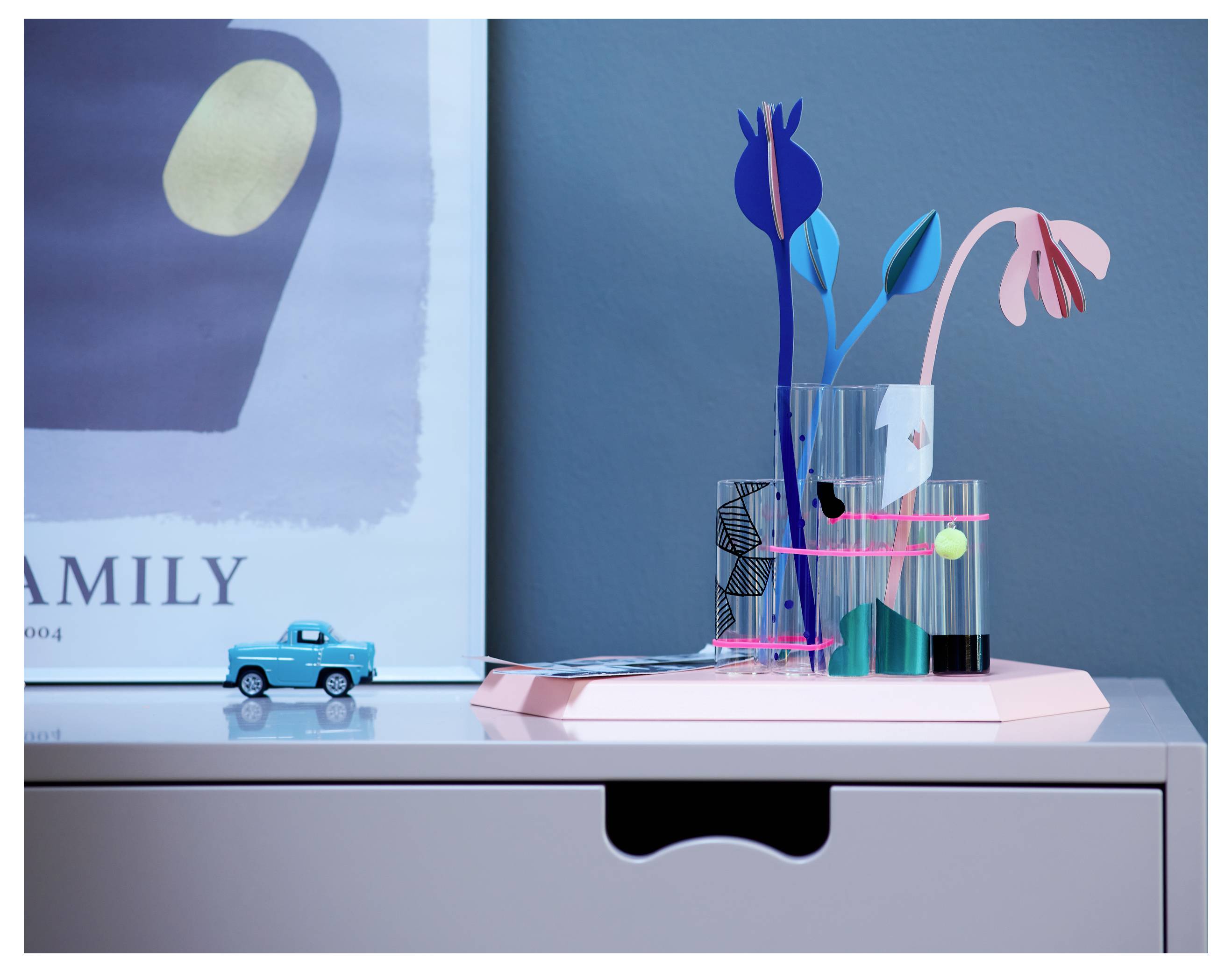 Kleiner blauer Spielzeuglastwagen neben einer geometrischen Blumenanordnung in Glas auf einem rosa Tablett auf einem Schrank, mit rahmenkunst im Hintergrund.