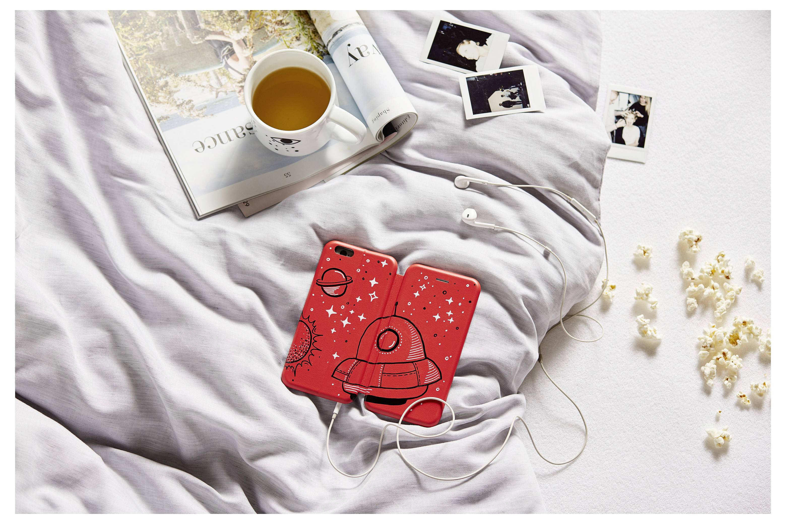 Rote Ohrhörer-Hülle mit weißem Kabel auf einem Bett neben einer Tasse Tee, einem Magazin und verstreuten Sofortbildfotos, was auf eine entspannte Hörsituation hindeutet.