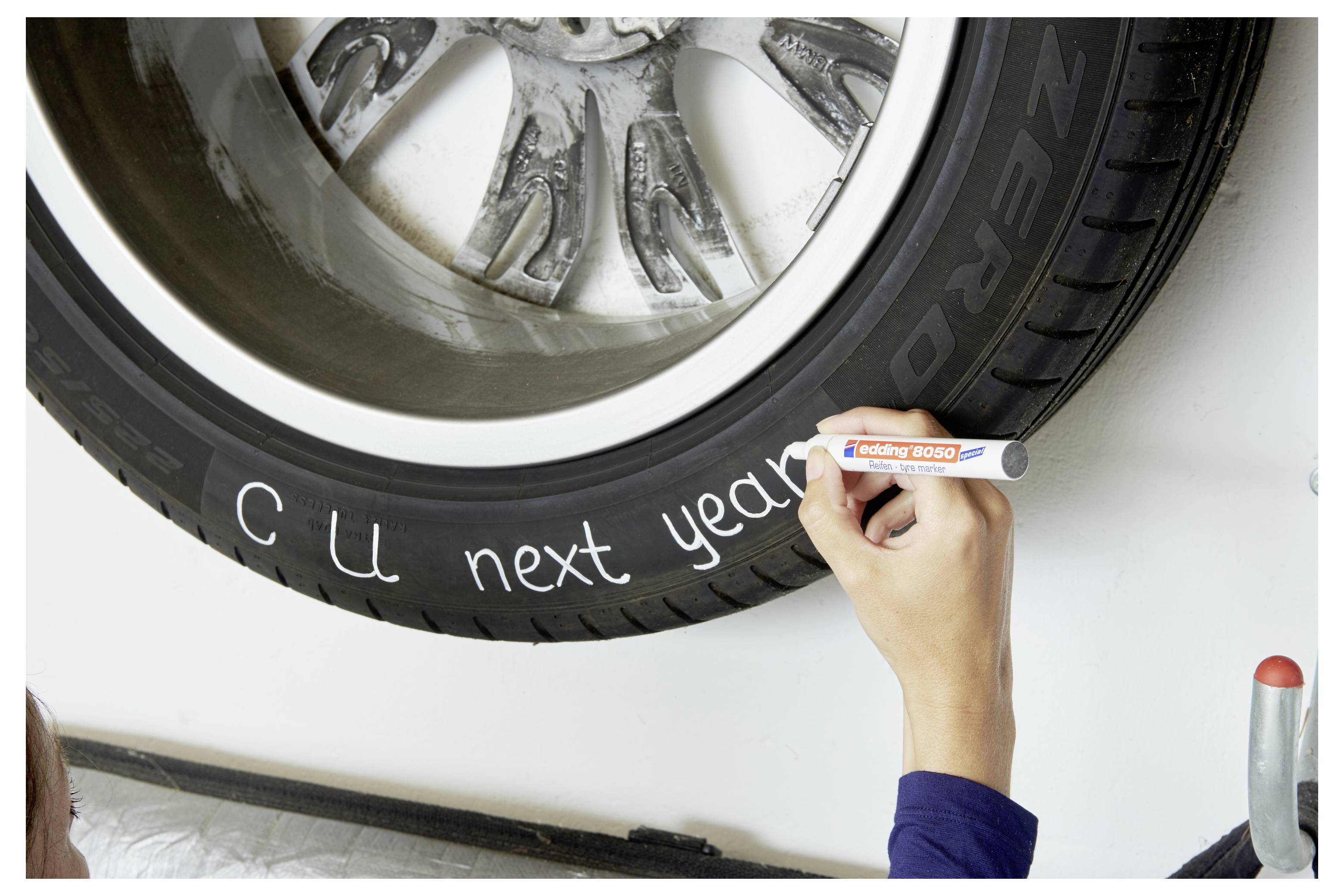 Eine Hand schreibt mit einem weißen Marker "CU next year" auf einen Autoreifen.