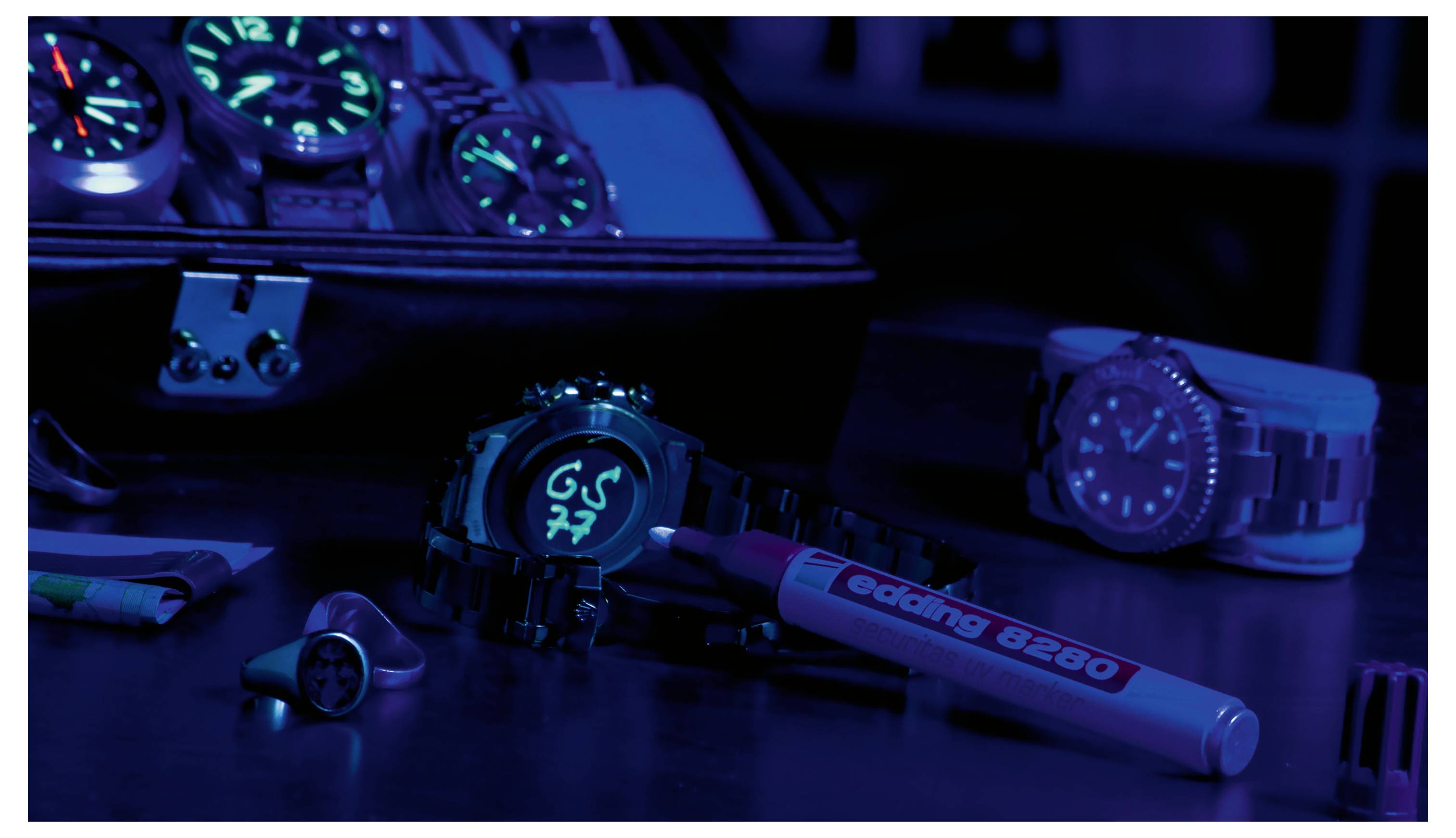 Verschiedene leuchtende Armbanduhren und Accessoires auf einem dunklen Tisch, beleuchtet in Blau und Violett, mit einem Marker im Vordergrund und einer Uhrenbox im Hintergrund.