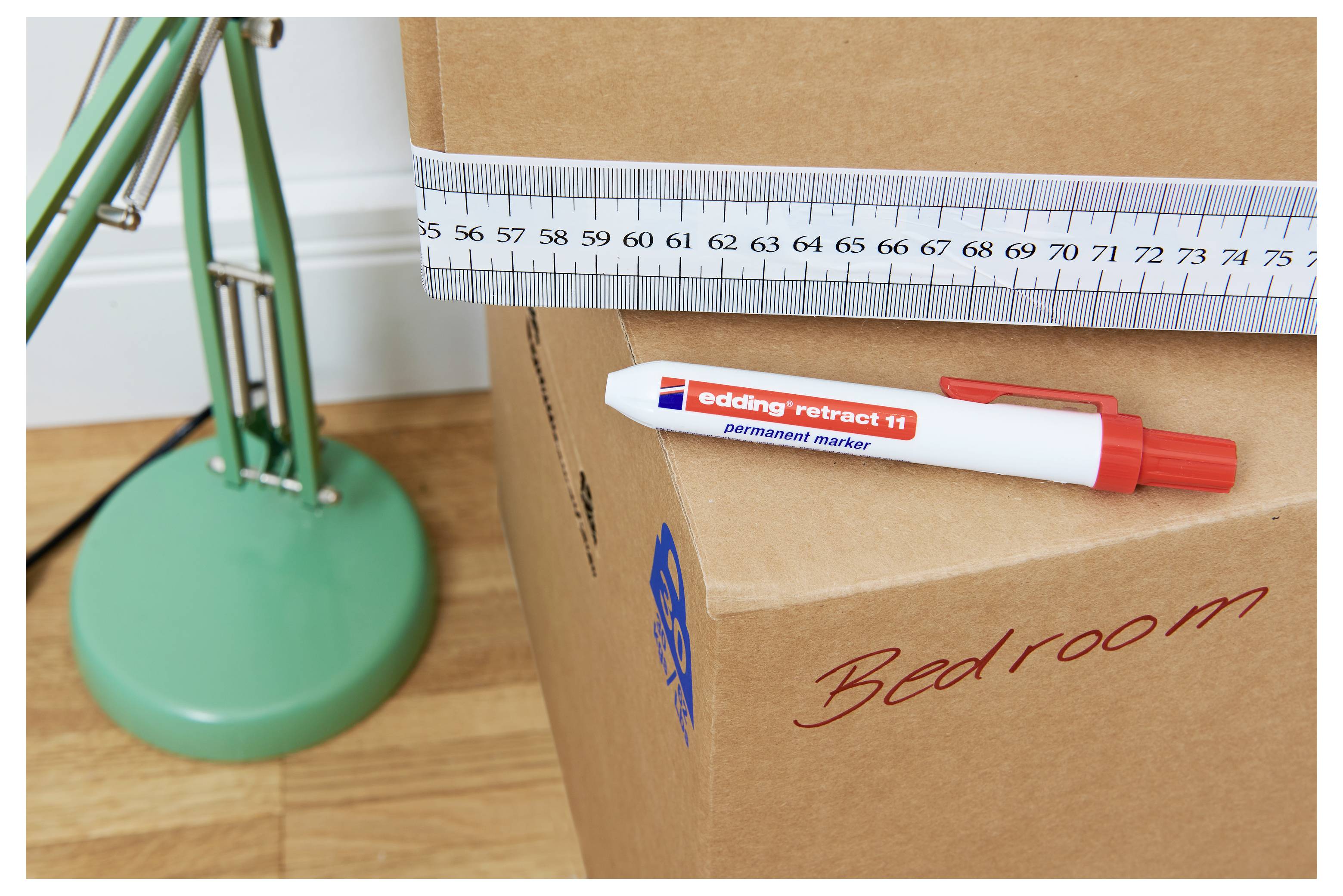 Karton-Umzugskarton mit der Aufschrift „Schlafzimmer" wird mit einem Stahllineal gemessen, mit einem Marker oben drauf und einer Lampenbase daneben.