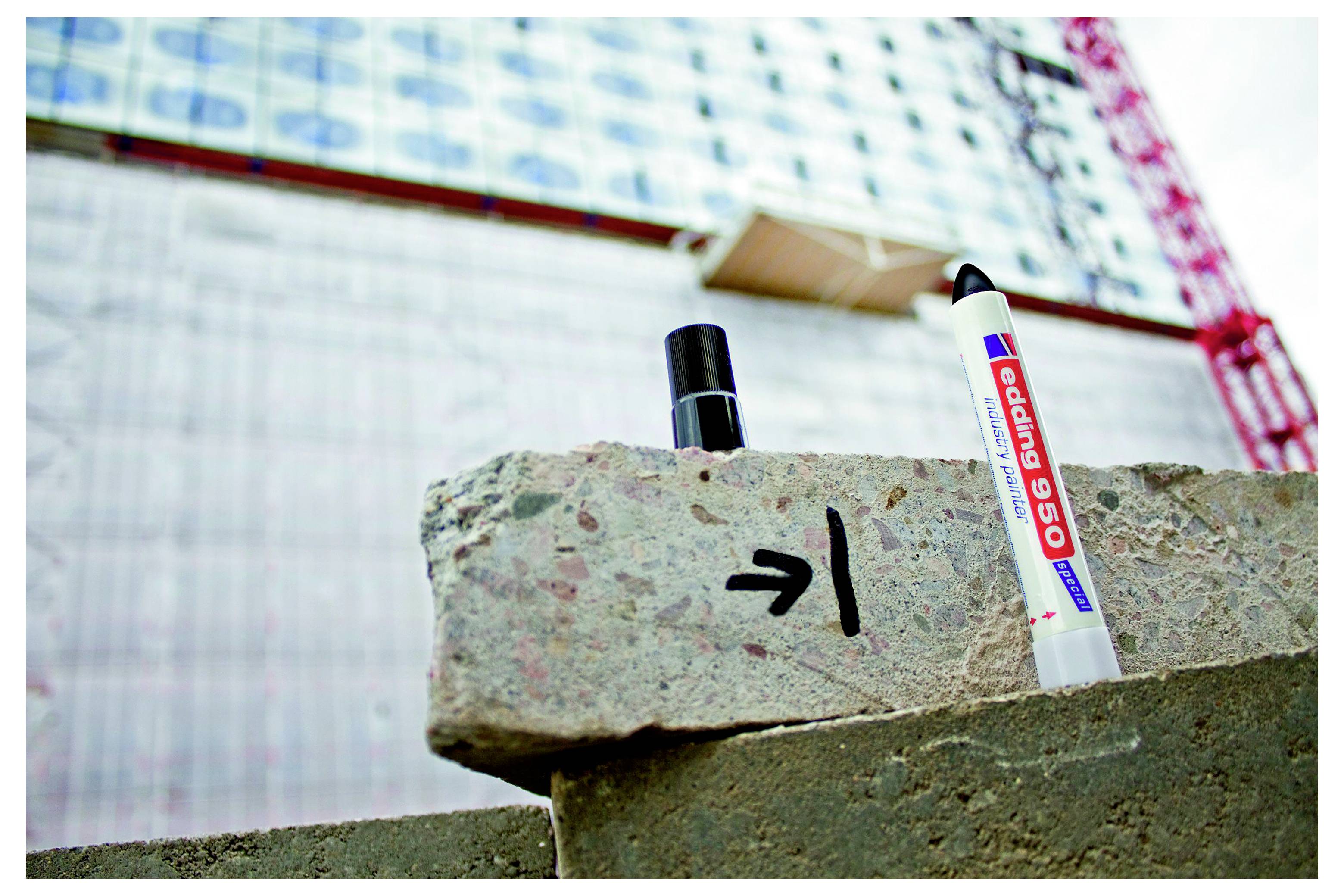 Betonblock mit Pfeil und Linie markiert, mit zwei Vermessungsmarken, vor einem Gerüst einer Baustelle