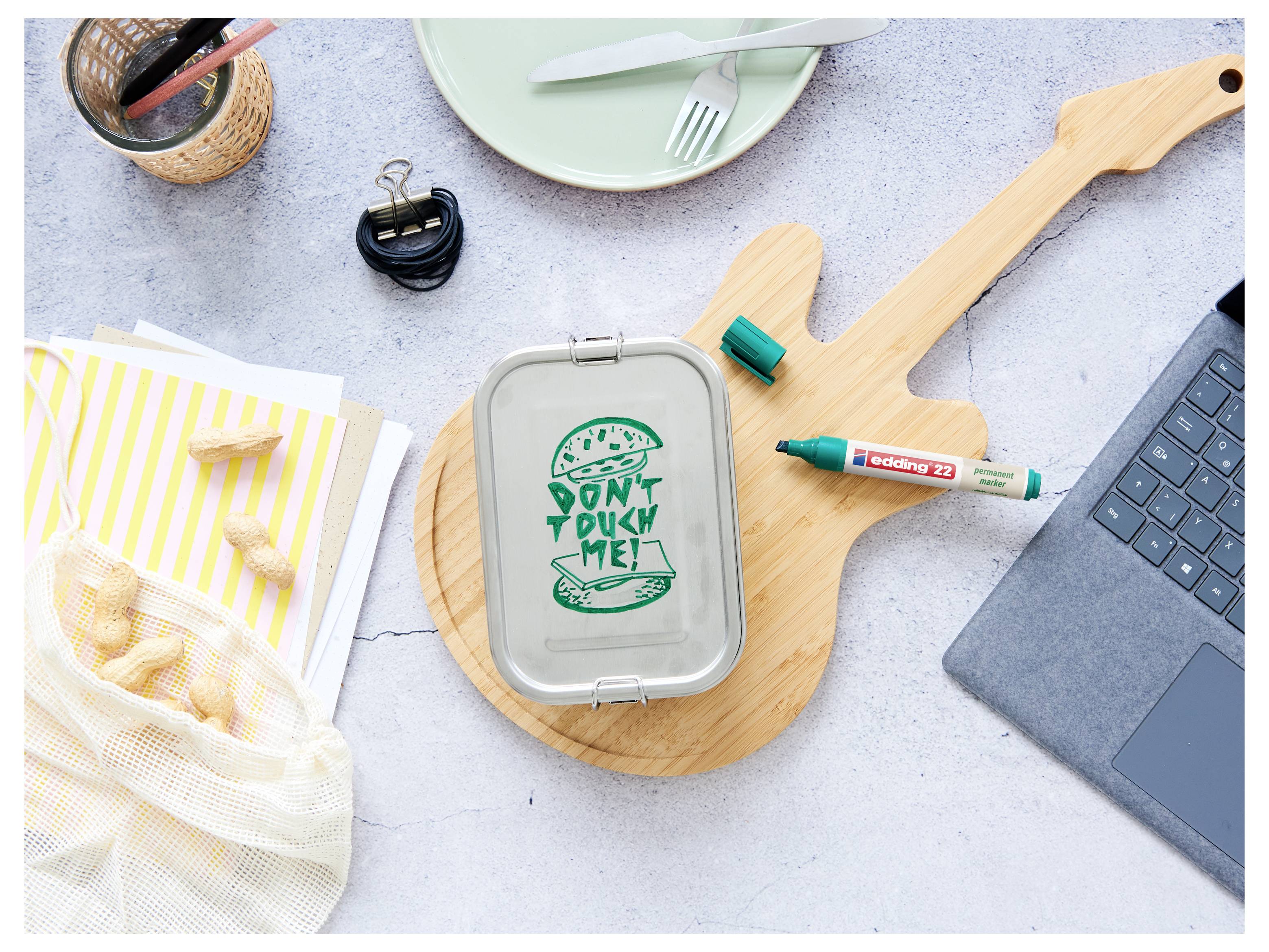 Kunststoff-Lunchbox mit der Aufschrift "MICH NICHT WEGWERFEN!", umgeben von wiederverwendbaren Gegenständen, einem Marker und einem Laptop, die eine Anti-Verschwendungs-Botschaft vermitteln.