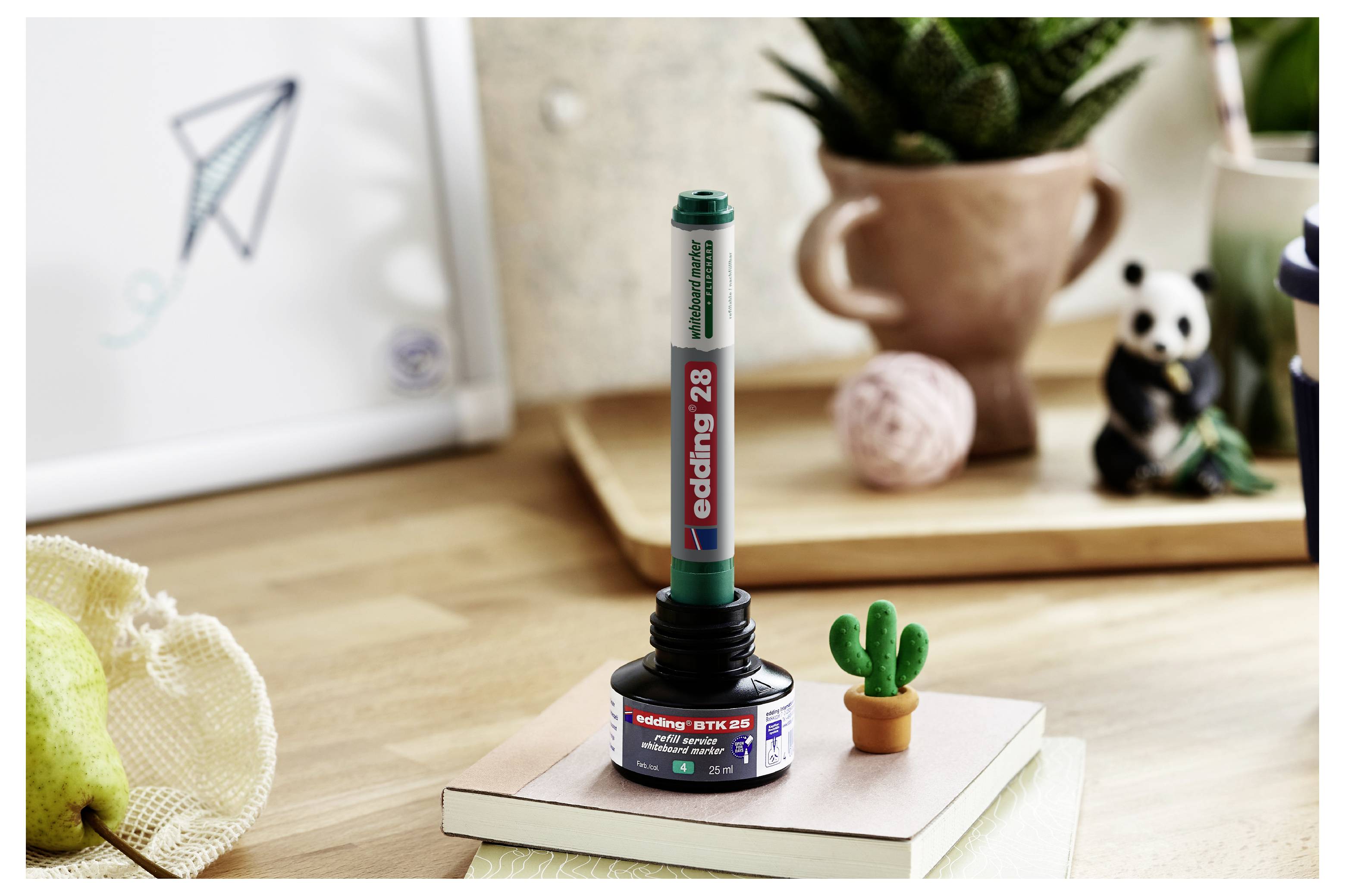 Ein grüner Edding-28-Marker steht aufrecht in einem schwarzen runden Halter auf einem Buch, auf einem Schreibtisch mit Topfpflanzen und einer gerahmten Zeichnung im Hintergrund.