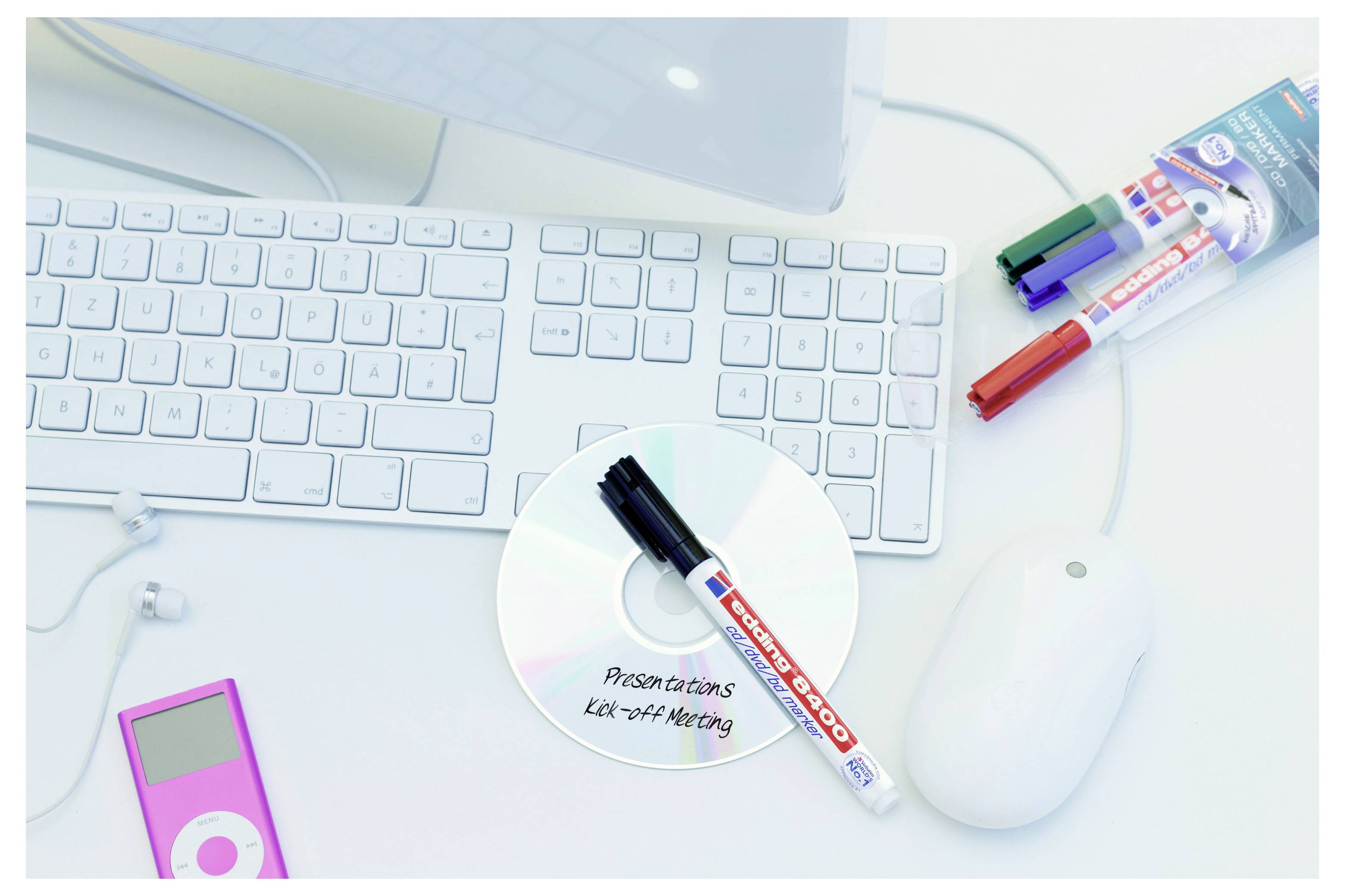 Draufsicht auf einen weißen Computertisch mit Tastatur, Maus, CD mit der Beschriftung „Presentations", Markierstiften und einem rosa MP3-Player.
