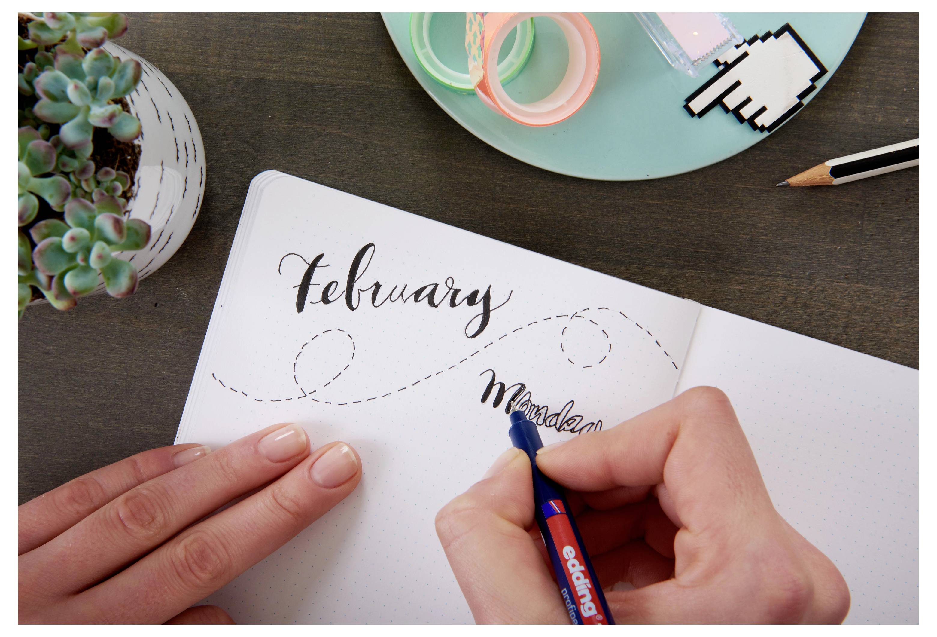 Hände schreiben "Februar" in ein Notizbuch mit Bleistifthilfe, was auf monatliche Bullet-Journal-Planung an einem Schreibtisch mit Schreibwaren hindeutet.