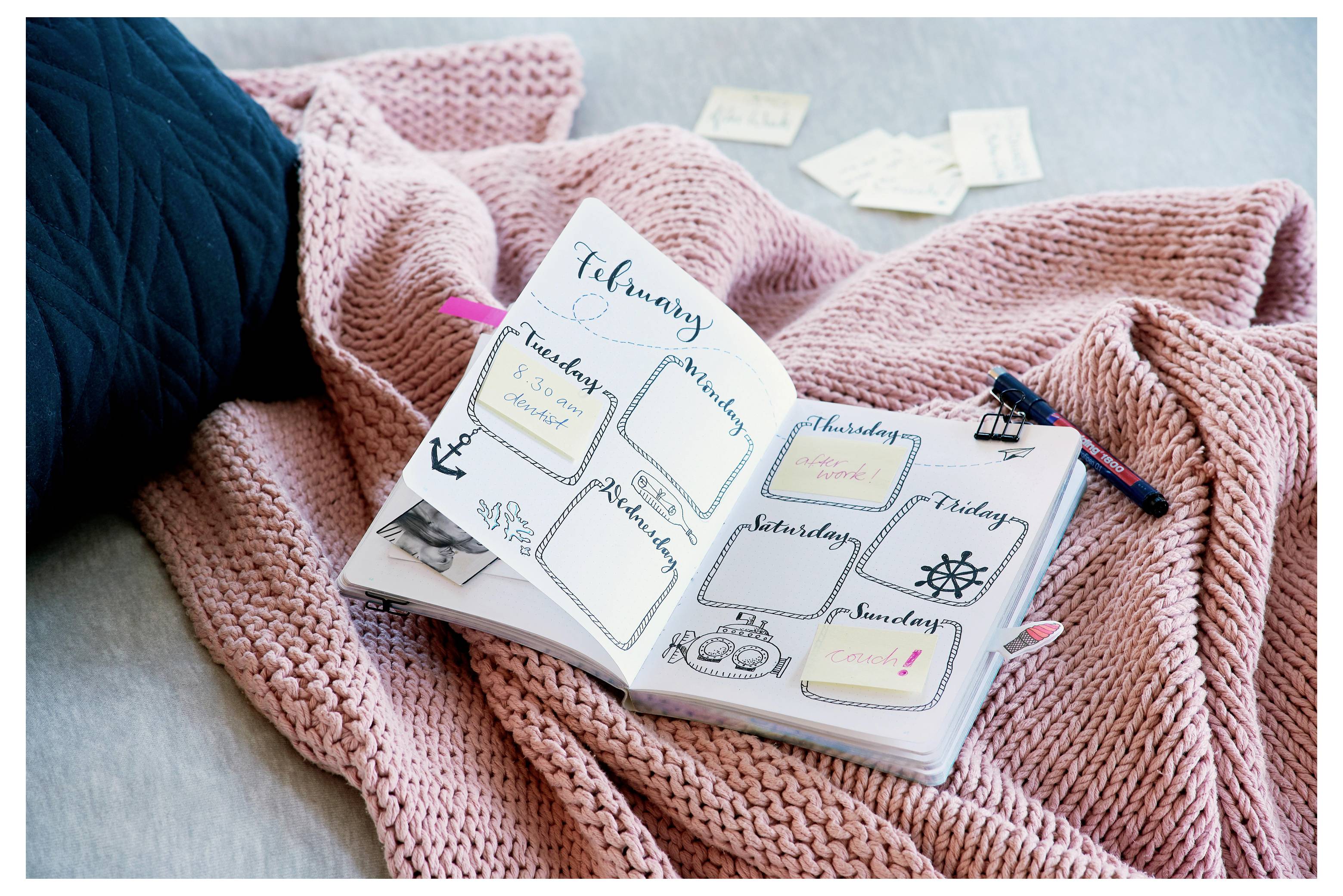 Ein offenes Bullet Journal auf einer rosa gestrickten Decke zeigt einen wöchentlichen Planer für Februar mit handgeschriebenen Tagesfeldern und dekorativen Symbolen.