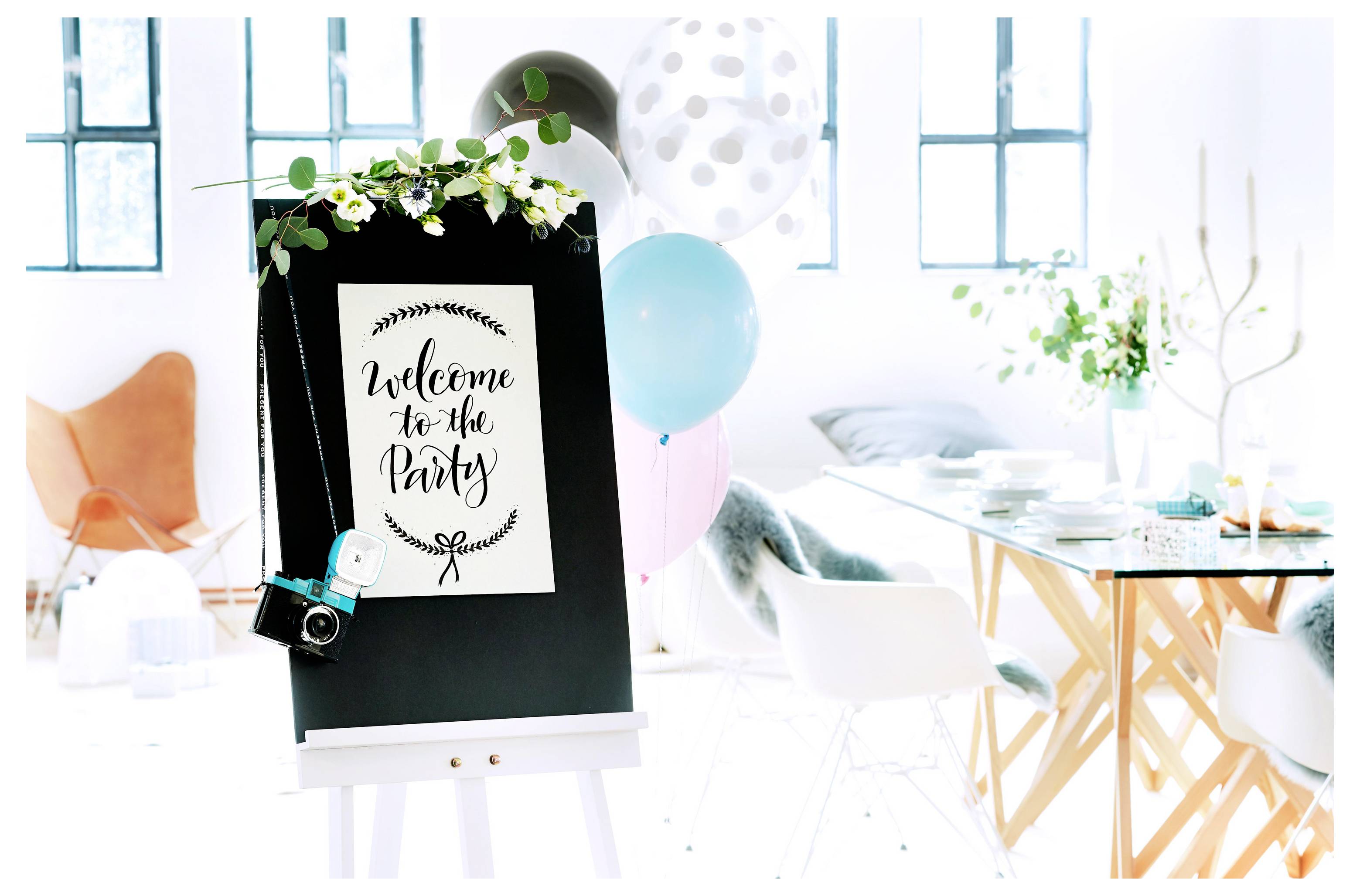 Willkommensschild zu einer Party auf einer Staffelei mit Blumen und Luftballons in einem hell dekorierten Raum mit Tischen und Stühlen.