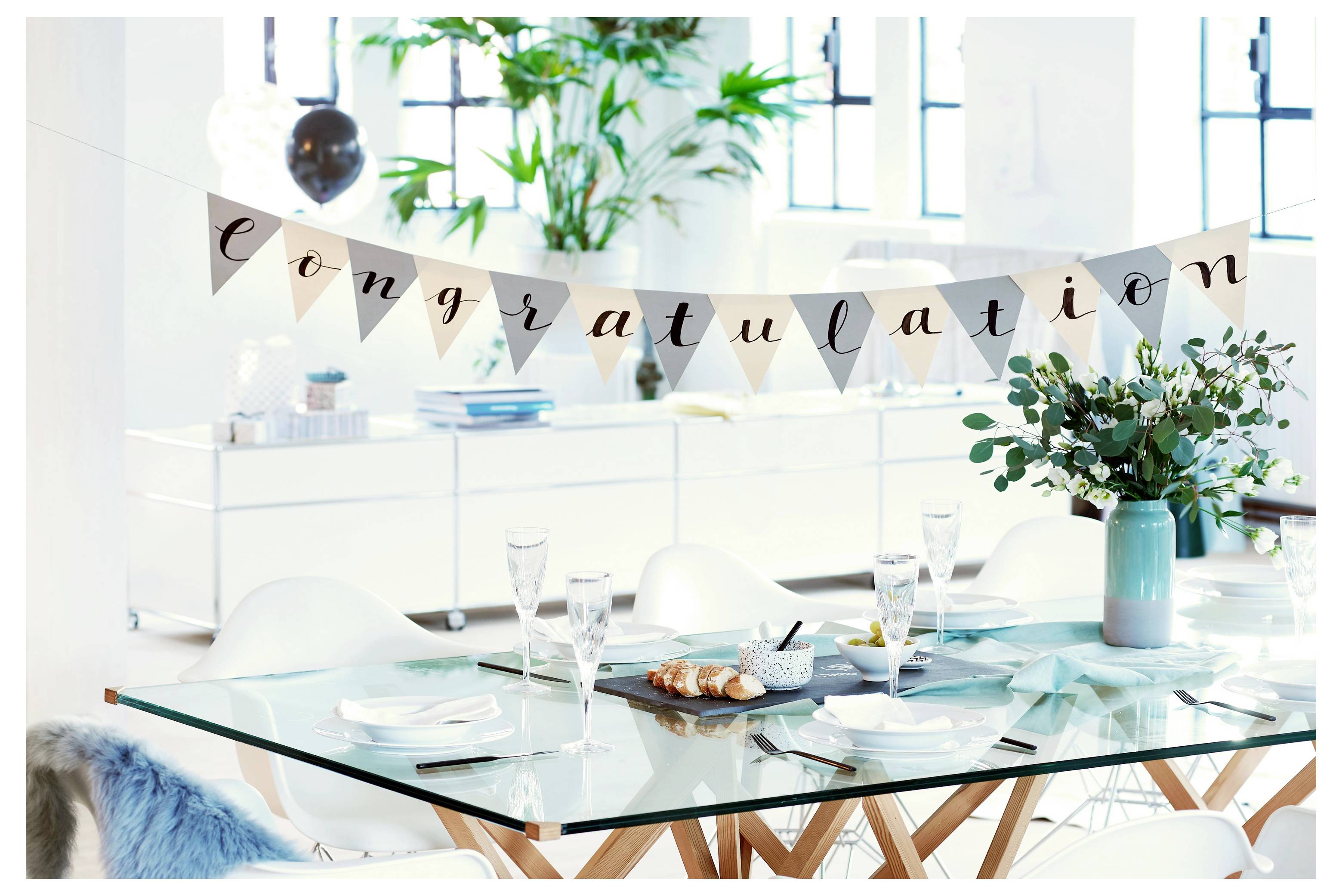 Helles Esszimmer, das für eine Feier dekoriert ist, mit einem "Glückwunsch"-Banner über einem gedeckten Tisch und Blumen in einer Vase.