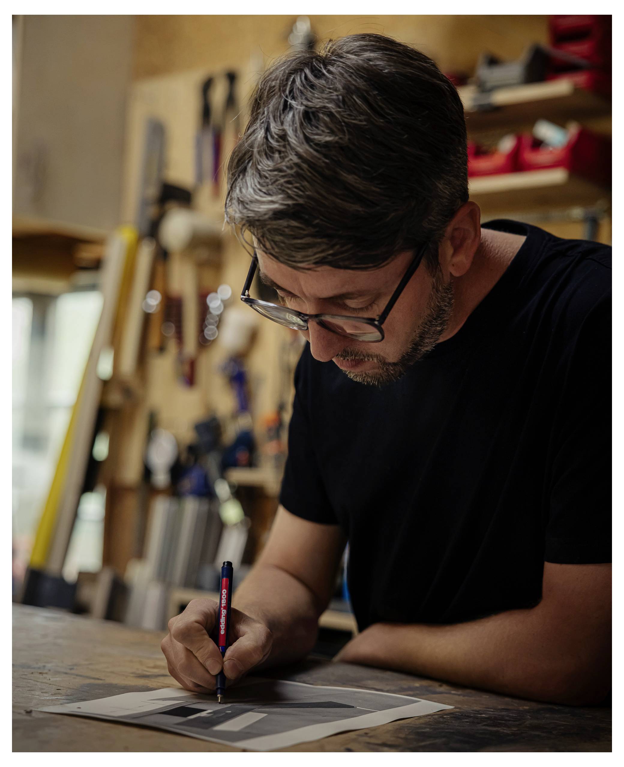 Ein Mann mit Brille lehnt sich über einen Arbeitstisch und skizziert auf Papier in einer unordentlichen Werkstatt oder einem Studio.
