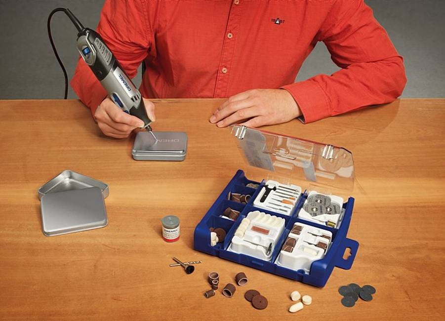 Eine Person in rotem Hemd graviert ein Metallkästchen mit einem Handwerkzeug. Auf dem Tisch liegen Kleinteile und ein Werkzeugset.