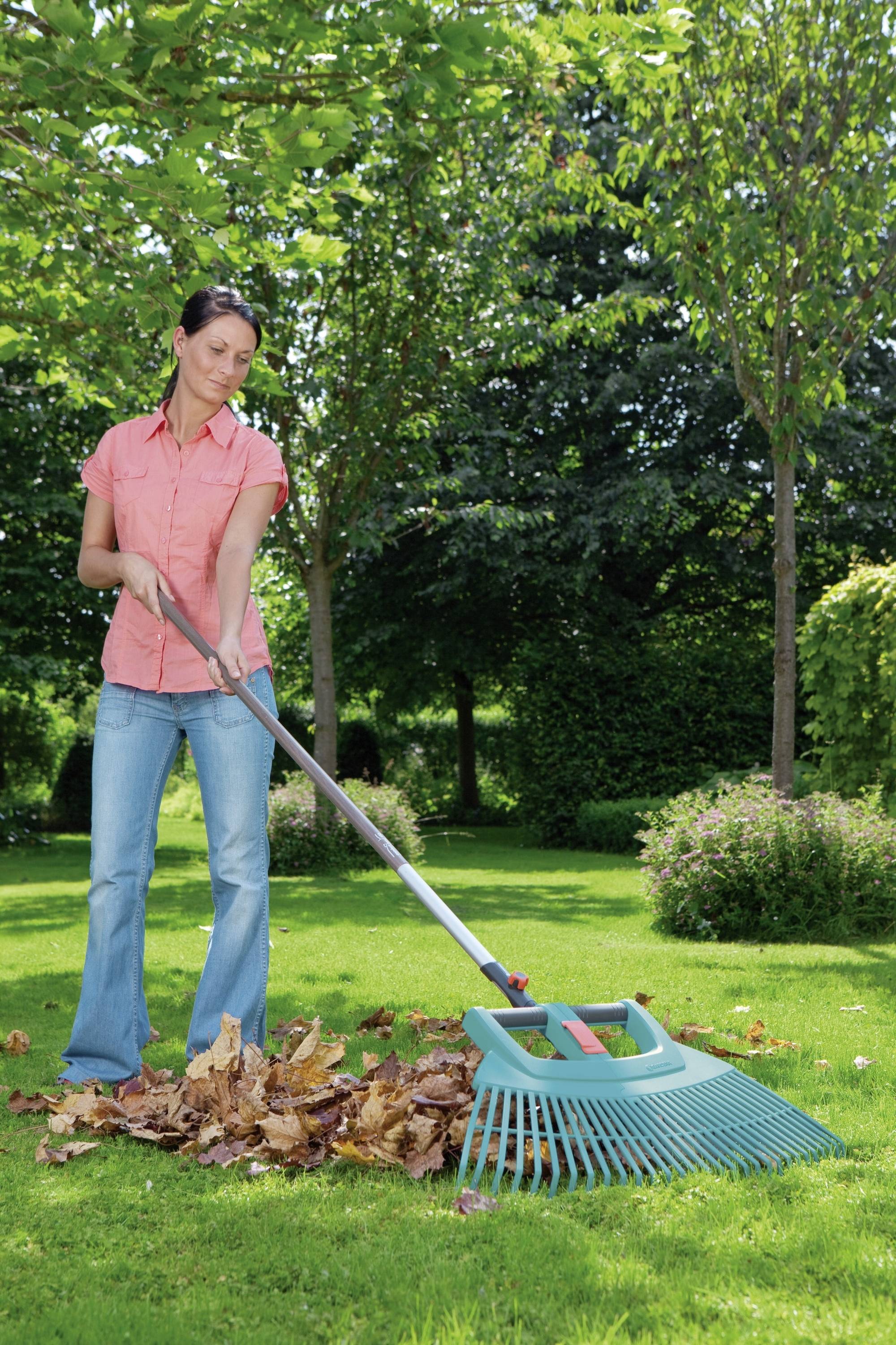 Frau im Garten harkt bunte Herbstblätter zusammen. Sie trägt ein rosa Hemd und blaue Jeans. Im Hintergrund sind grüne Bäume zu sehen.