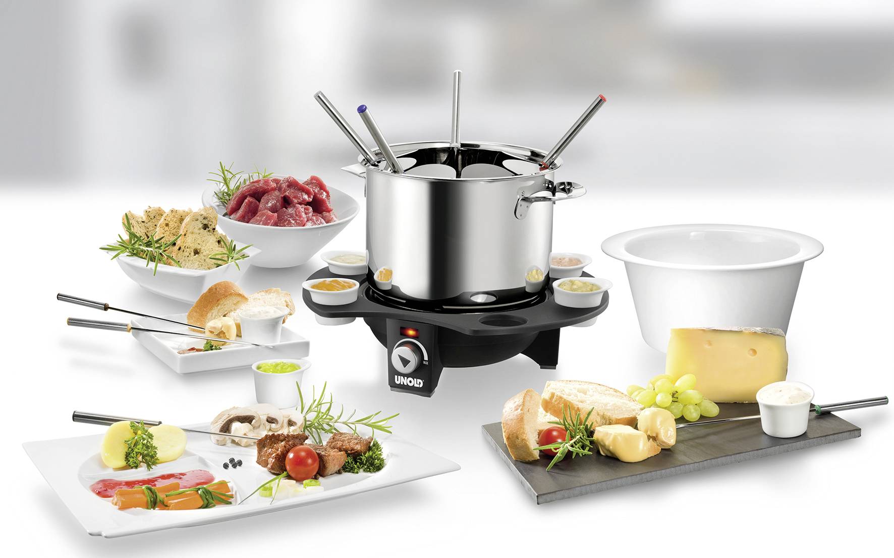 Ein Fondueset mit acht Gabeln, umgeben von Fleisch, Brot, Käse, Trauben und Saucen auf Tellern.