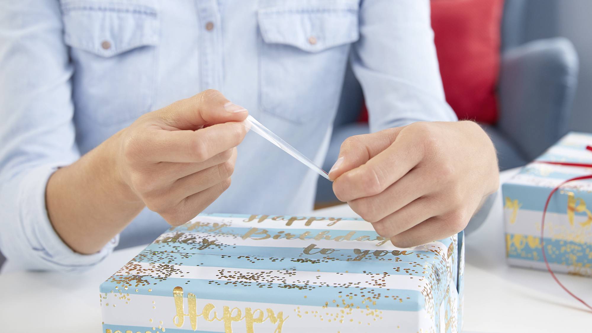 Eine Person verpackt ein Geschenk mit blauem, weißem und goldenem Papier, das 'Happy Birthday to you' sagt.