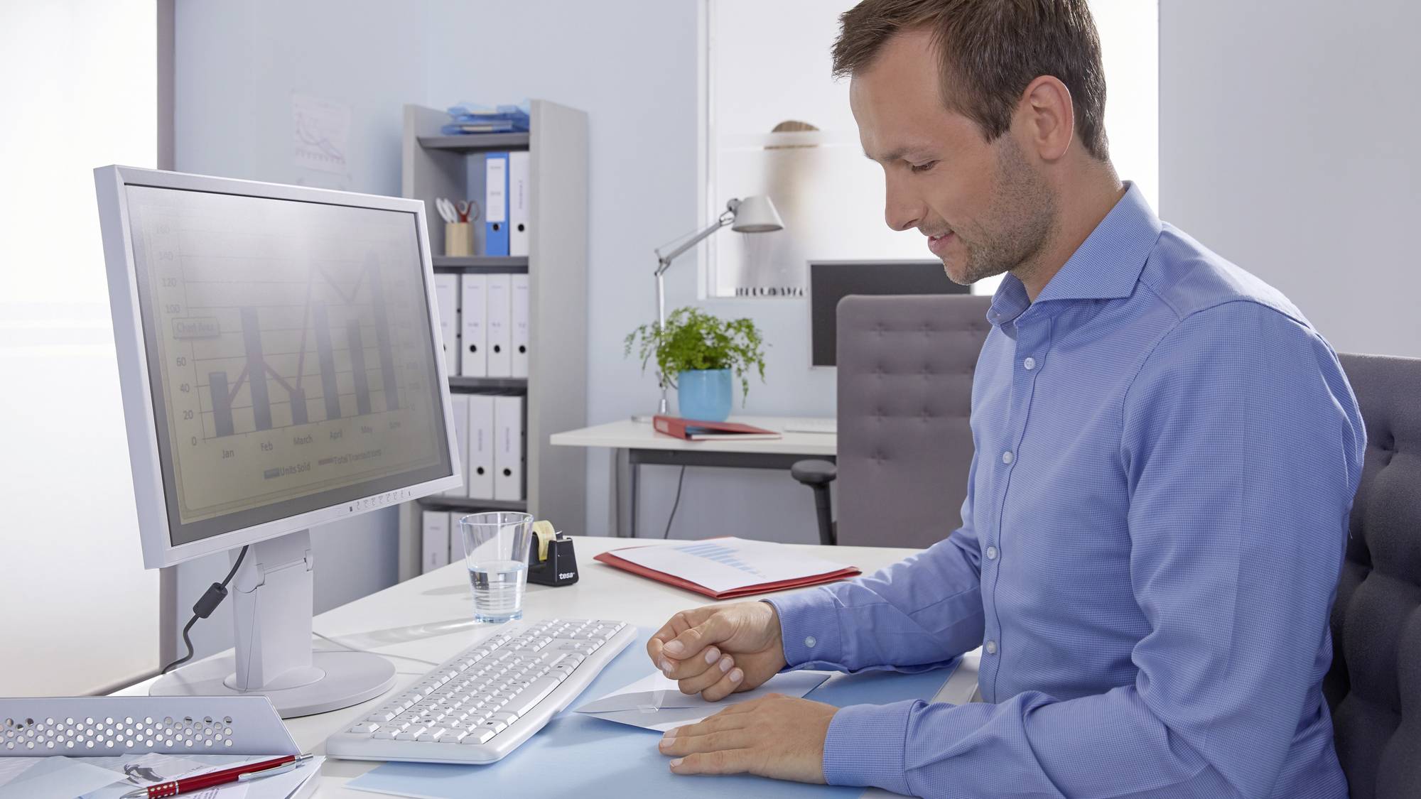 Ein Mann in einem Büro arbeitet an Dokumenten am Schreibtisch neben einem Computerbildschirm mit Diagrammen. Im Hintergrund Regale.