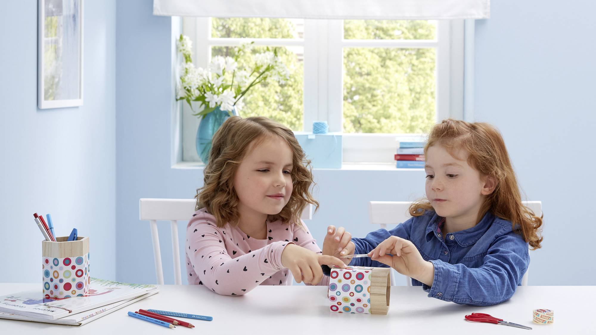 Zwei Kinder sitzen an einem Tisch in einem hellen Raum und basteln gemeinsam. Auf dem Tisch liegen Buntstifte und Bastelmaterial.