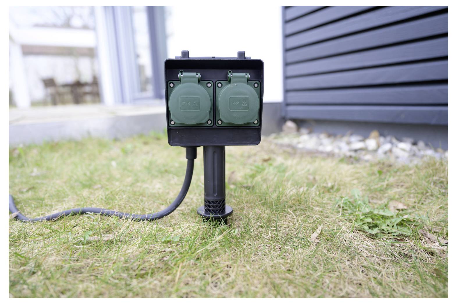 Outdoor-Doppelsteckdosenkastens auf einem kurzen Pfosten im Gras neben einem Haus montiert, mit einem Kabel, das von links angeschlossen ist.