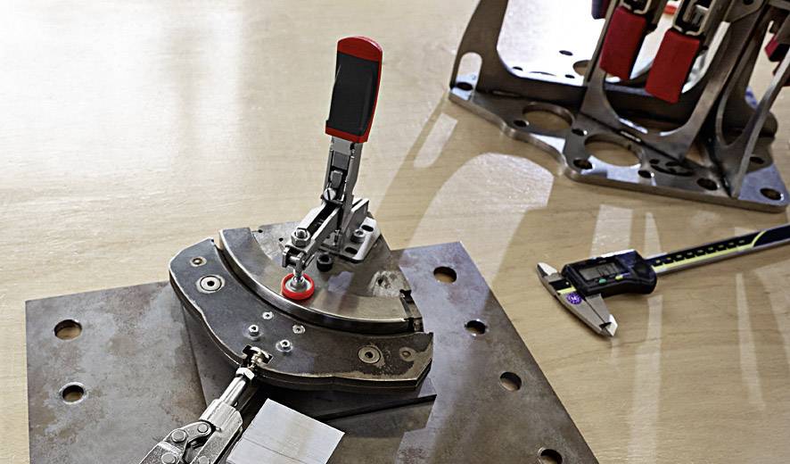 Eine Metallplatte mit Halteklemmen und einem Winkelmesser auf einem Holztisch, rechts ein Schieblehre, dient zur präzisen Messung.