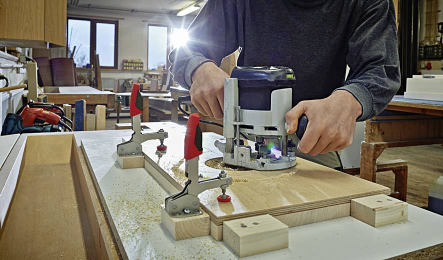 Eine Person benutzt eine Oberfräse, um Holz auf einem Werkbank in einer Werkstatt zu bearbeiten. Holzteile sind mit Klammern fixiert.