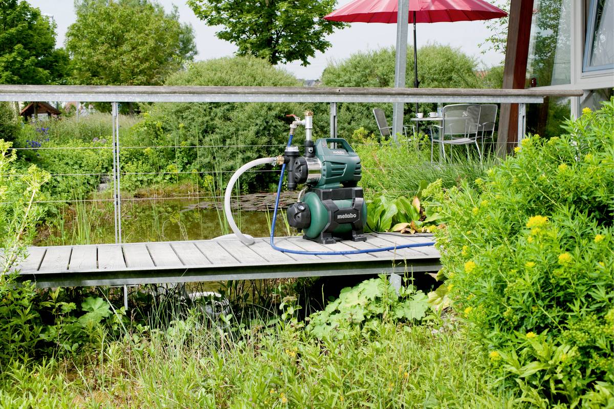 Eine grüne Wasserpumpe steht auf einem Holzsteg vor einem Teich, umgeben von üppigen Pflanzen und Sträuchern im Garten.