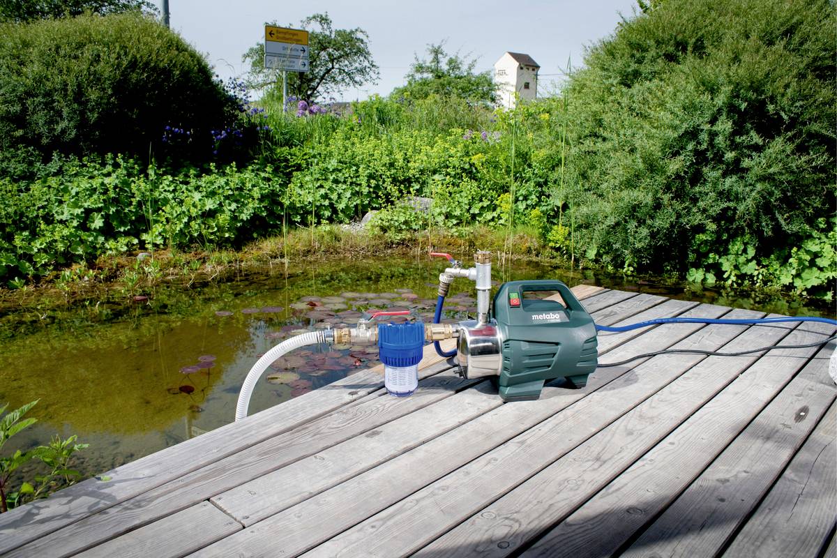 Eine grüne Wasserpumpe mit Schlauch steht auf einer Holzterrasse, neben einem Teich mit Seerosen und umgeben von dichter Vegetation.