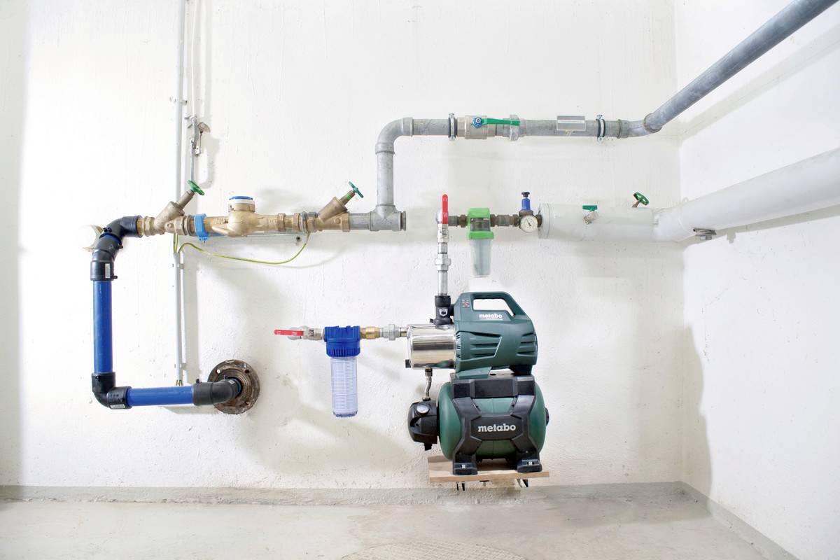 Wasserpumpe mit blauen und grauen Rohrleitungen an einer weißen Wand, verschiedene Ventile zur Kontrolle des Wasserflusses.