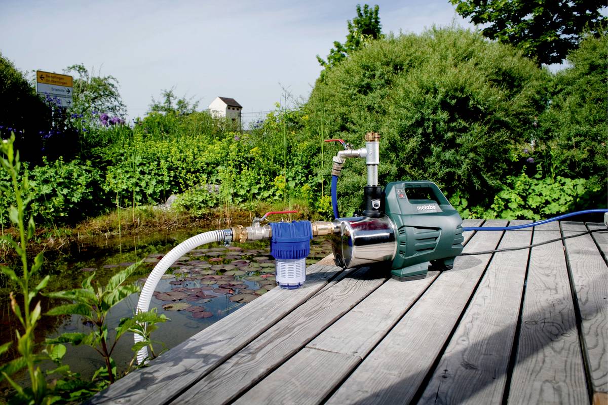 Eine Wasserpumpe auf einer Holzterrasse neben einem Teich, umgeben von grünen Pflanzen. Die Pumpe hat ein blaues Filtersystem.