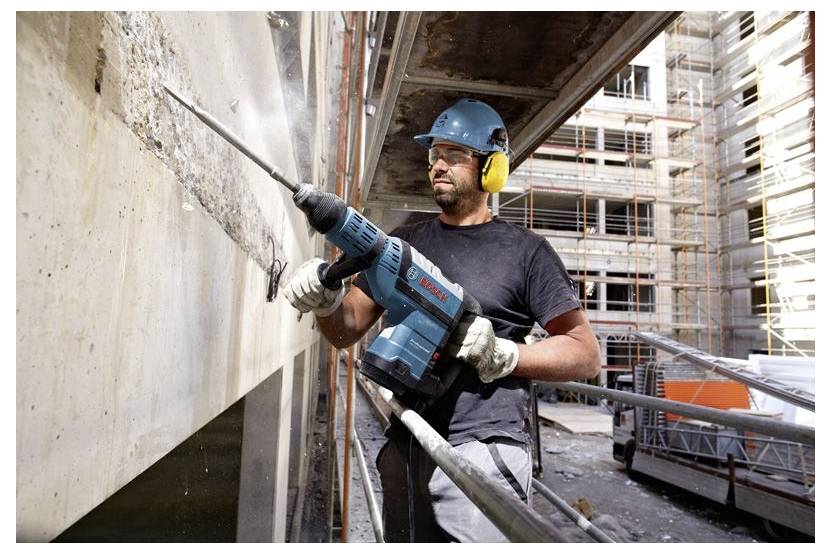 Ein Bauarbeiter in Sicherheitsausrüstung benutzt einen Bohrhammer an einer Betonwand auf einer Baustelle, umgeben von Gerüsten und Baumaterialien.
