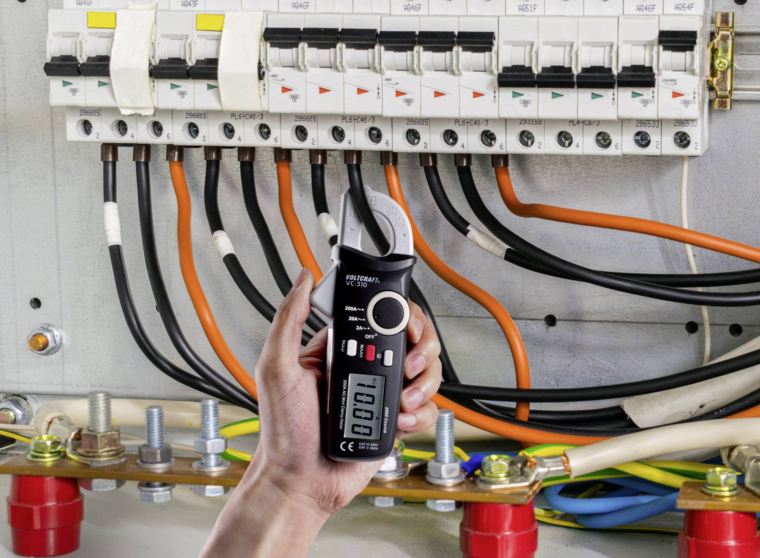 Eine Hand hält ein schwarzes Strommessgerät vor einer geöffneten Schalttafel mit mehreren Kabeln und Sicherungsschaltern.
